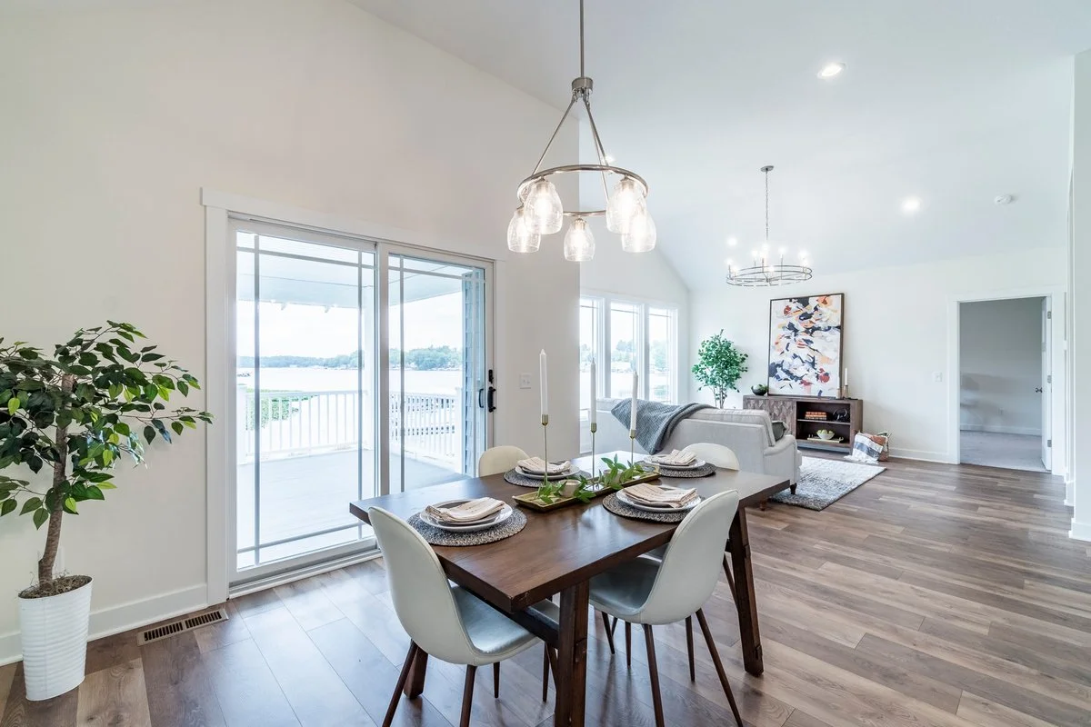 Open-concept living and dining area with a wooden dining table set for four, overlooking a balcony with a view of a body of water, natural light, white walls, and modern decor.