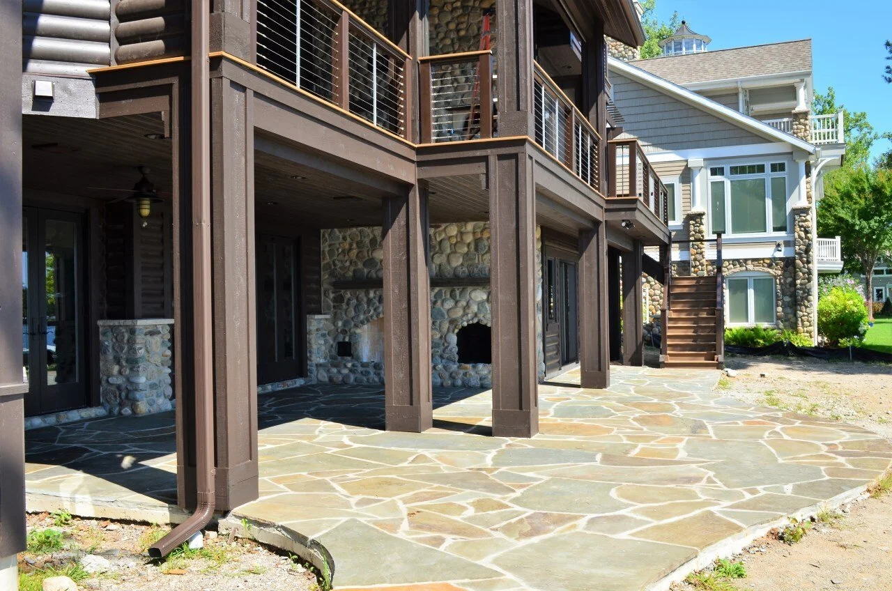 View of the back of a house with a stone patio, porch, and stairs leading to an upper deck, featuring stone and wood exterior with multiple windows and balconies.