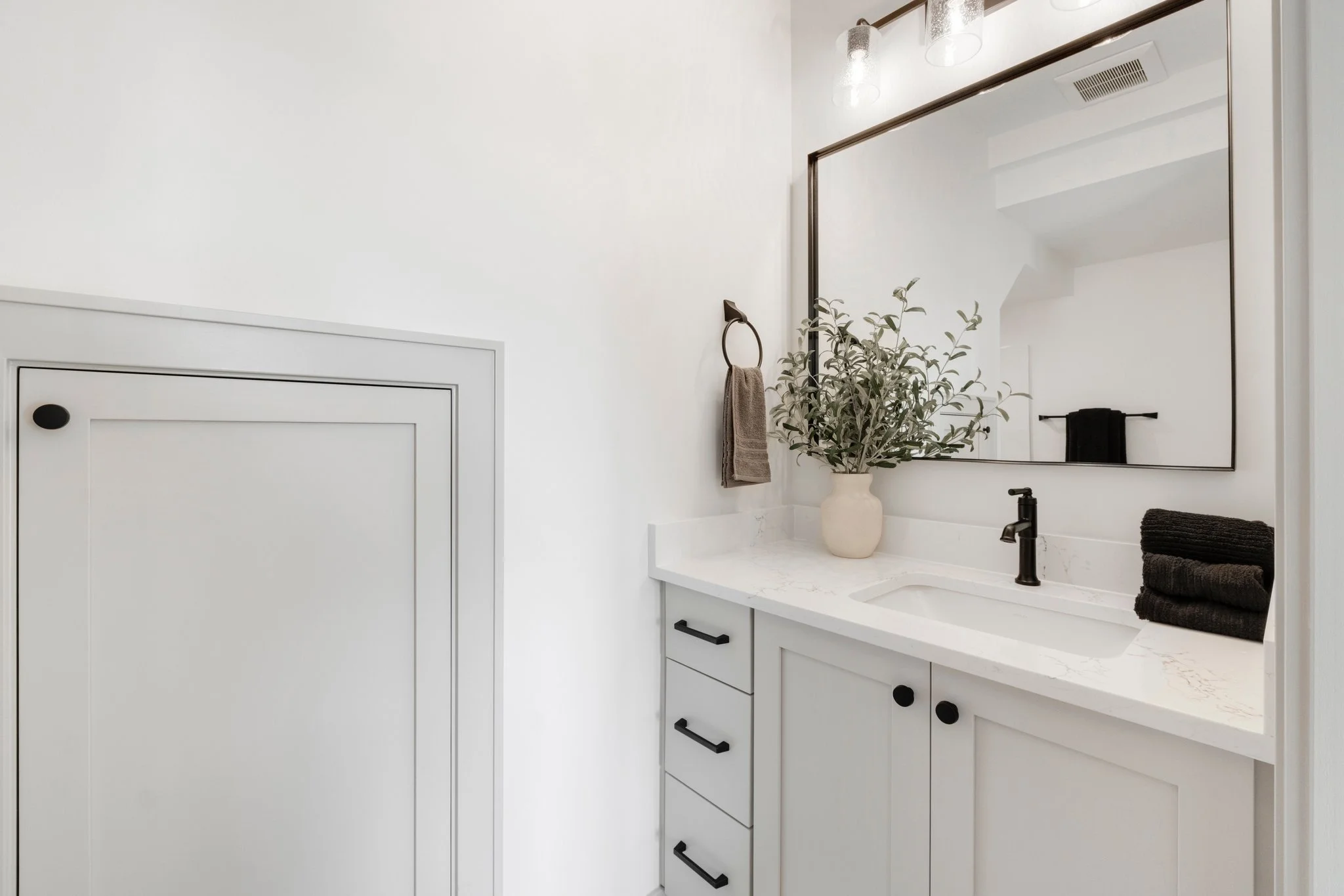 Bathroom vanity with white cabinet, black hardware, marble countertop, black faucet, large mirror, potted plant, beige towel, and black towels, with a white wall and closed cabinet door to the left.