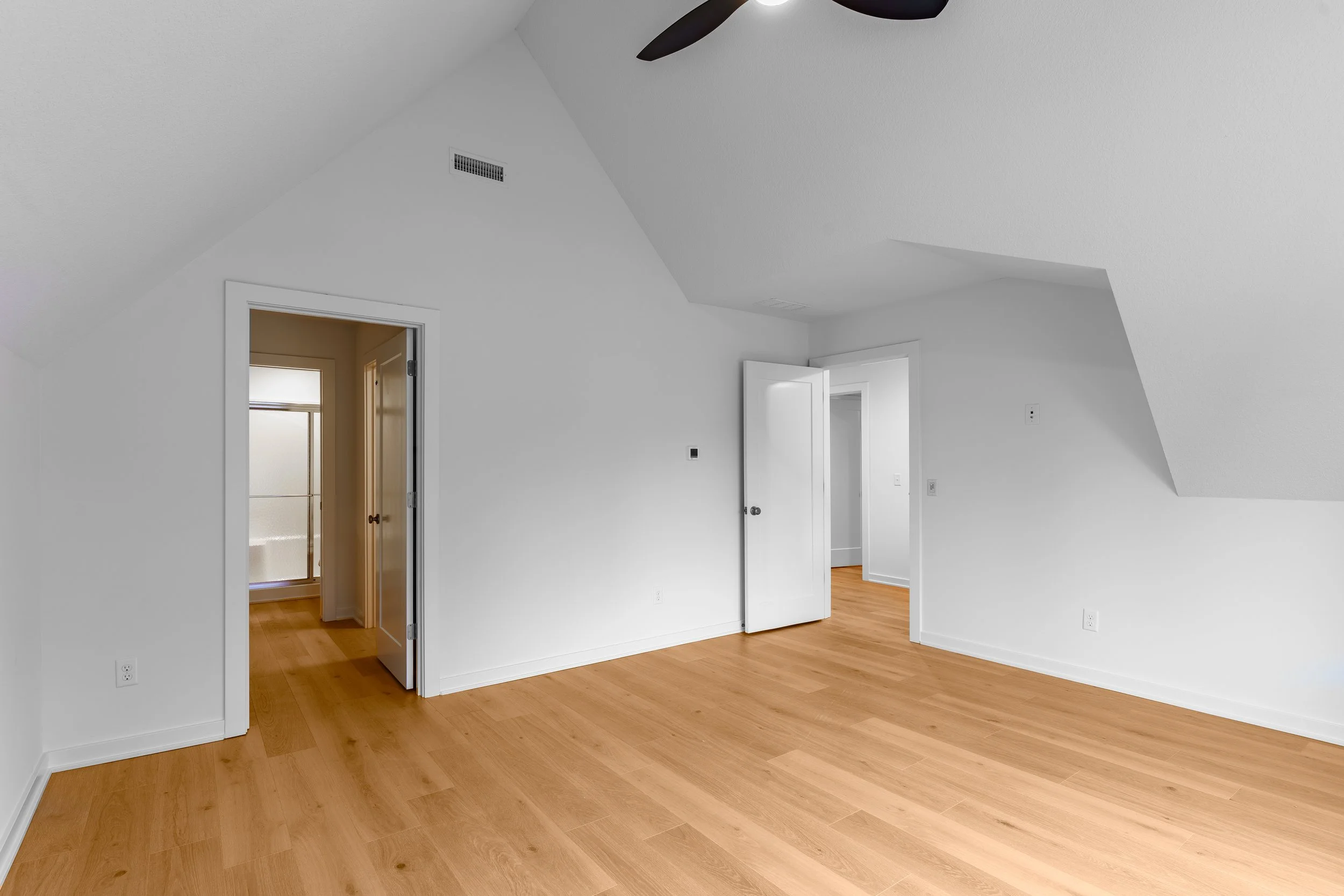Empty room with white walls, wooden floor, ceiling fan, and multiple open doors leading to other rooms.