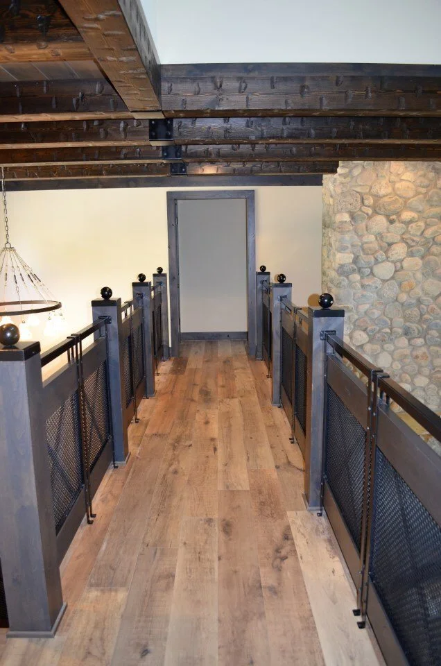 A hallway with wooden flooring, dark metal railing with black ball caps, stone wall on the right, exposed wooden beams on the ceiling, and a closed beige door at the end.