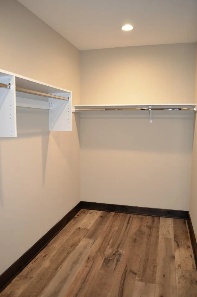 Empty walk-in closet with white shelves and hanging rods, beige walls, wooden floor, and recessed ceiling light.