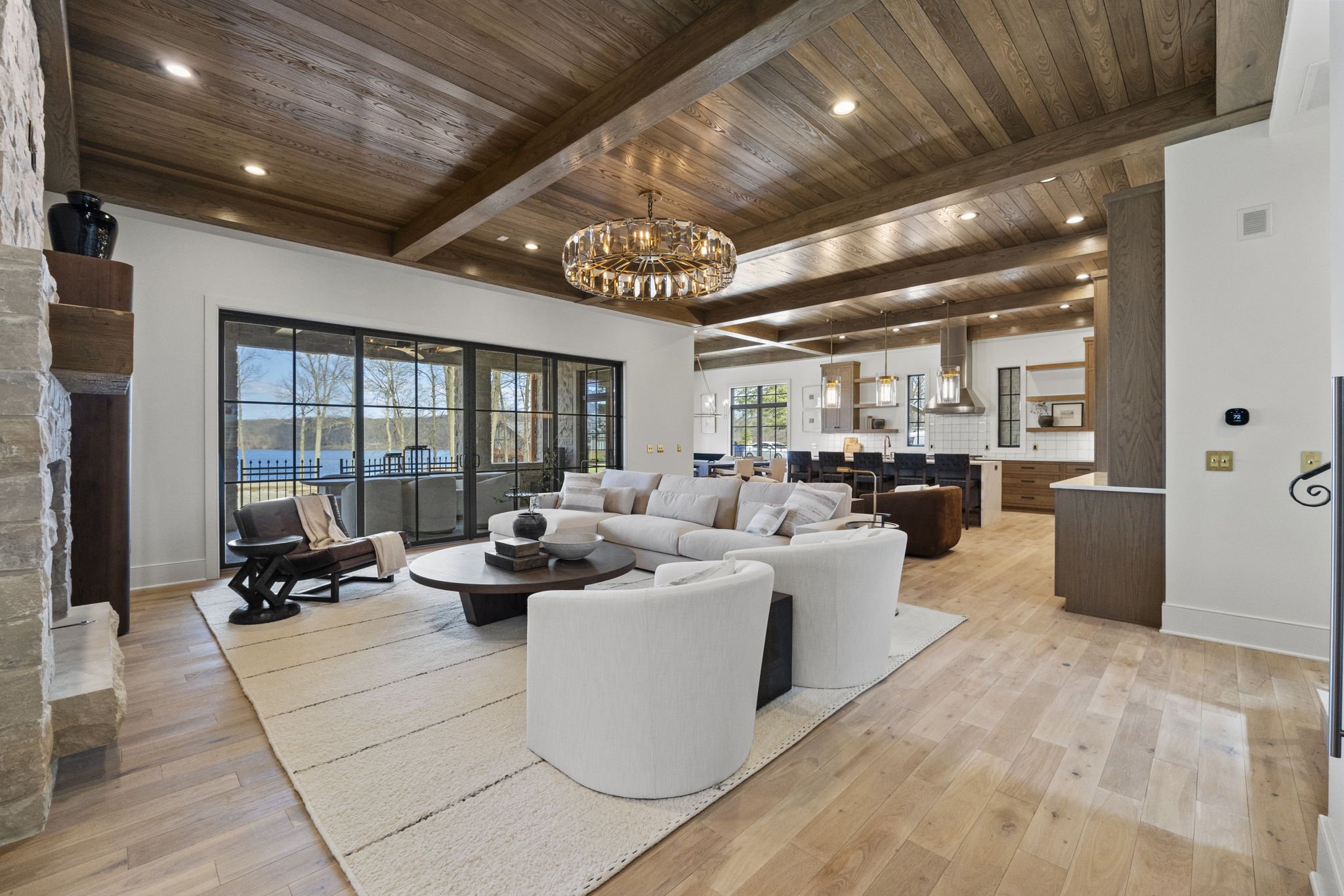 Modern open-plan living room with wooden ceiling, white and beige furniture, large sliding glass doors leading outside, visible kitchen area in the background, and a view of a lake and trees through the windows.
