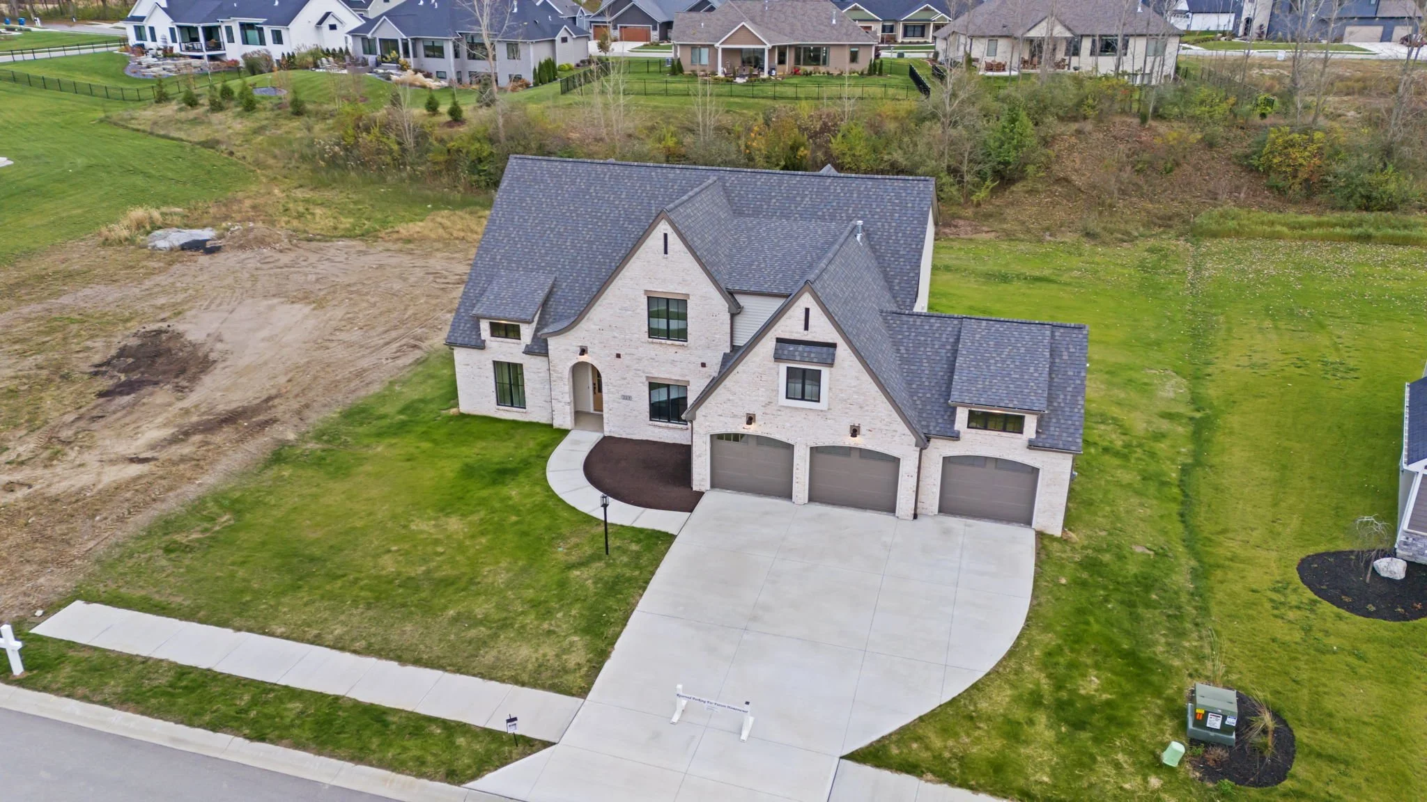 An aerial view of a new two-story house with a three-car garage, gray roof, brick exterior, and a lawn with a curved sidewalk and street in a suburban neighborhood.