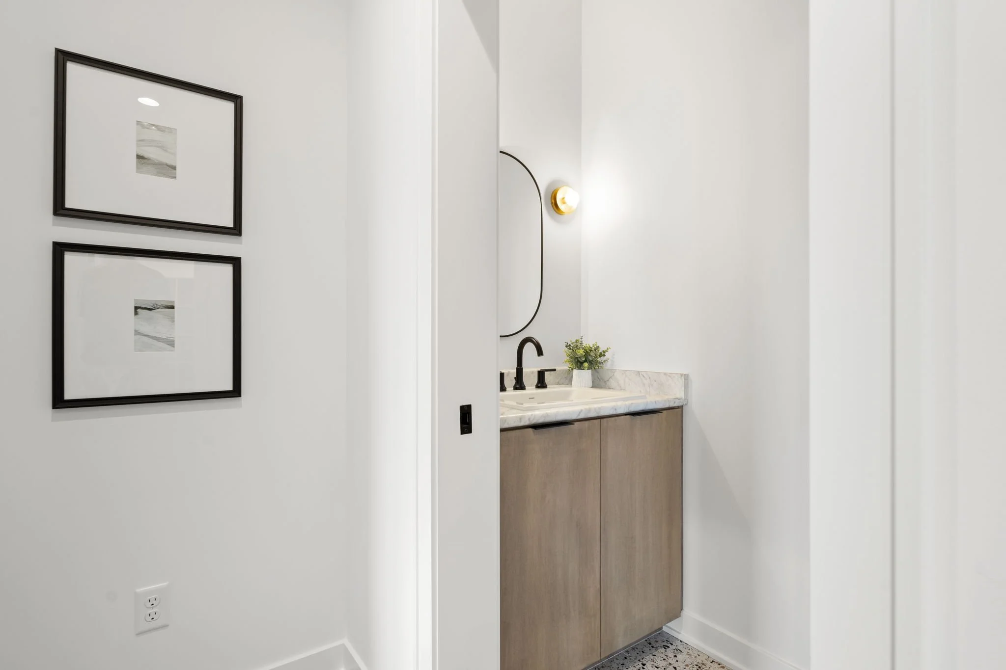 A minimalist bathroom with a white wall, two black-framed landscape pictures, a small black-framed mirror, a modern wall sconce, light wood vanity with a marble countertop, black faucet, and a small potted plant.