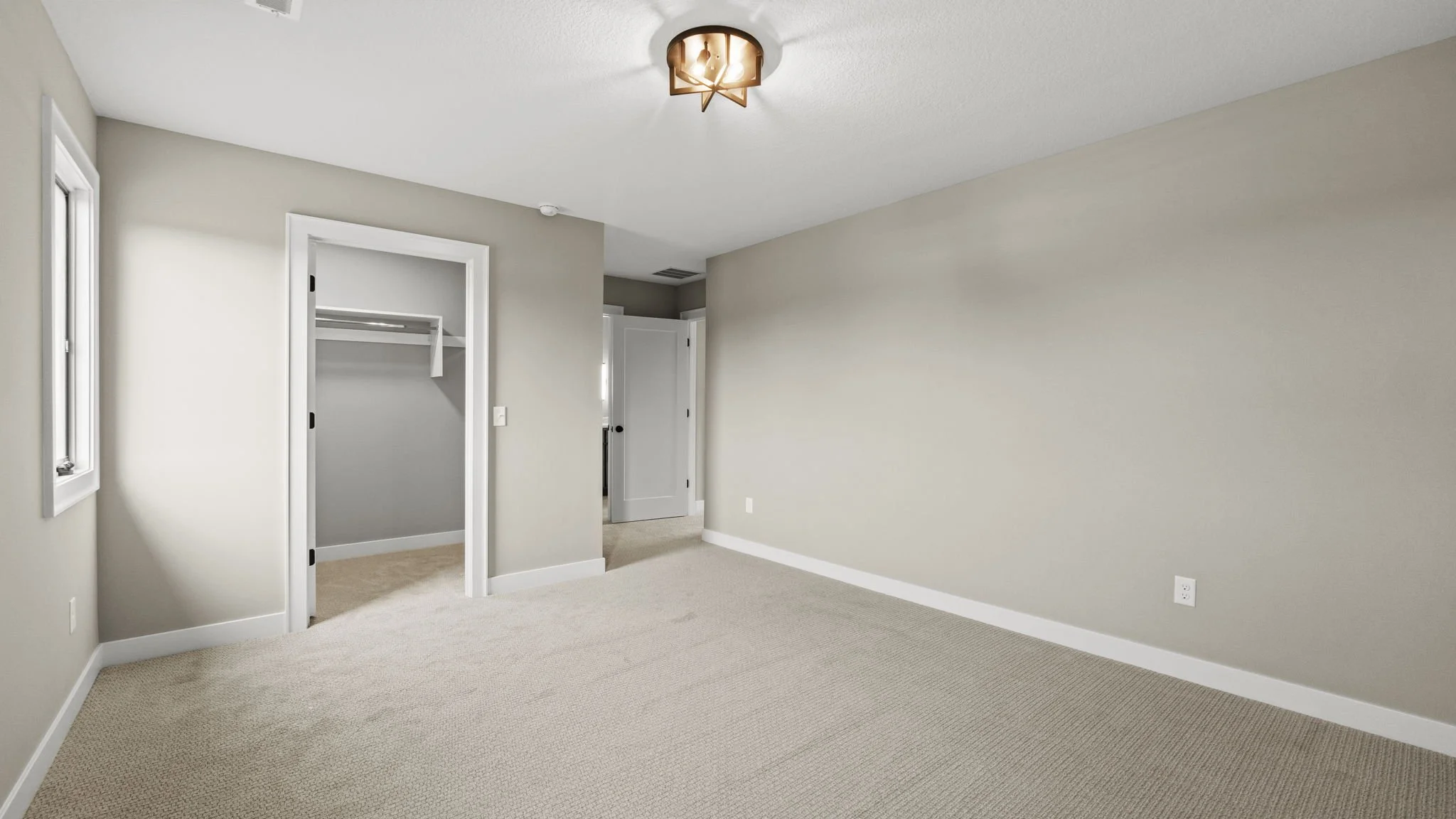 Empty bedroom with beige carpet, light gray walls, white door, closet, window, and ceiling light fixture.