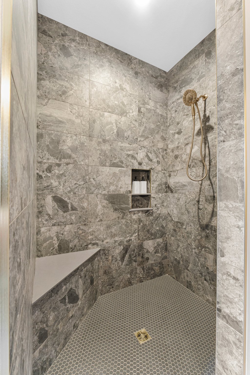 A modern shower with gray marble tiles, a built-in corner shelf with shampoo and body wash, a gold-colored handheld showerhead, and small hexagonal gray tiles on the shower floor.