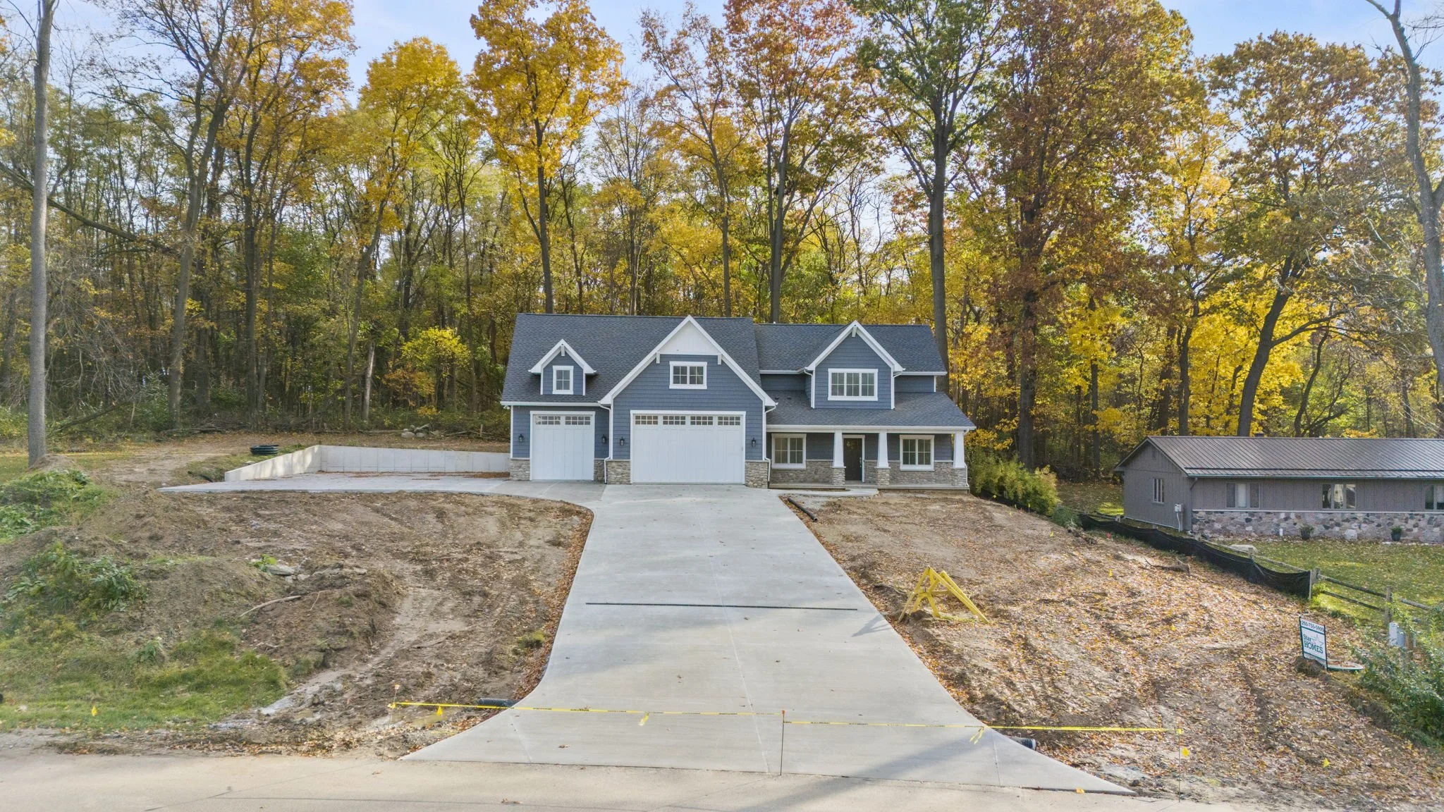 Newly built blue house with white trim and attached garage, surrounded by trees with fall foliage, and a concrete driveway in the front yard.