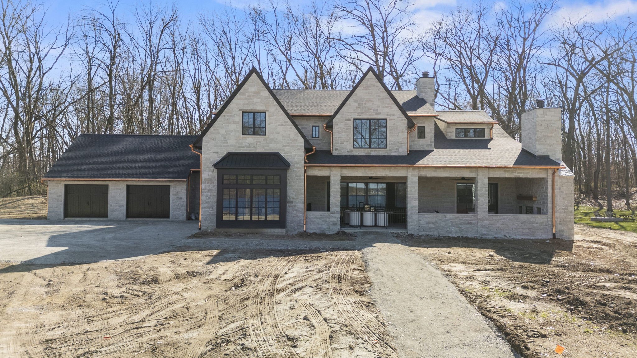 Under construction house with a brick facade, a front porch, and multiple gabled roofs, surrounded by bare trees and a dirt driveway.