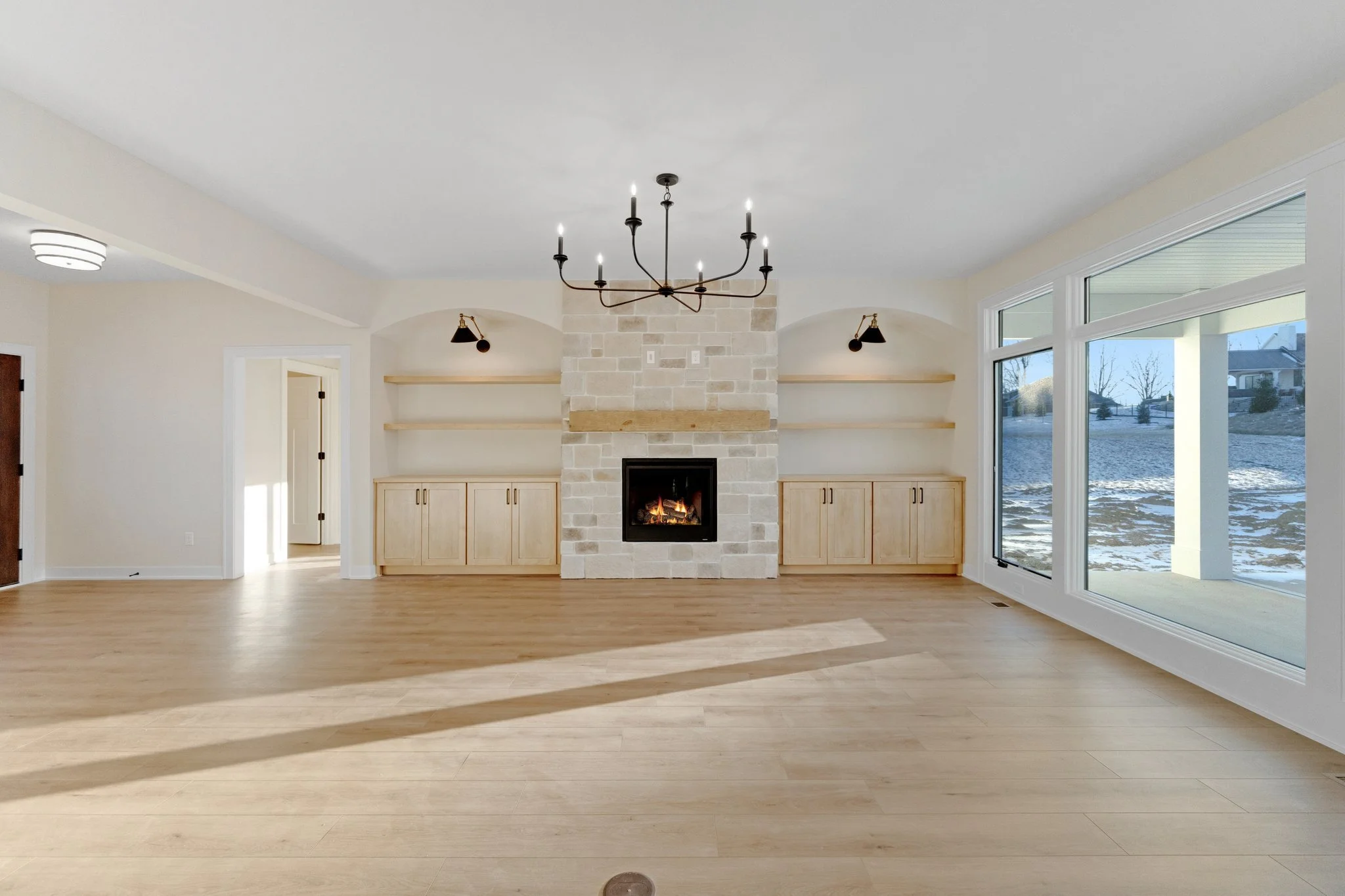 Empty living room with light wood flooring, a stone fireplace, built-in cabinets, large windows showing an outdoor patio and snow-covered yard, and a black chandelier hanging from the ceiling.