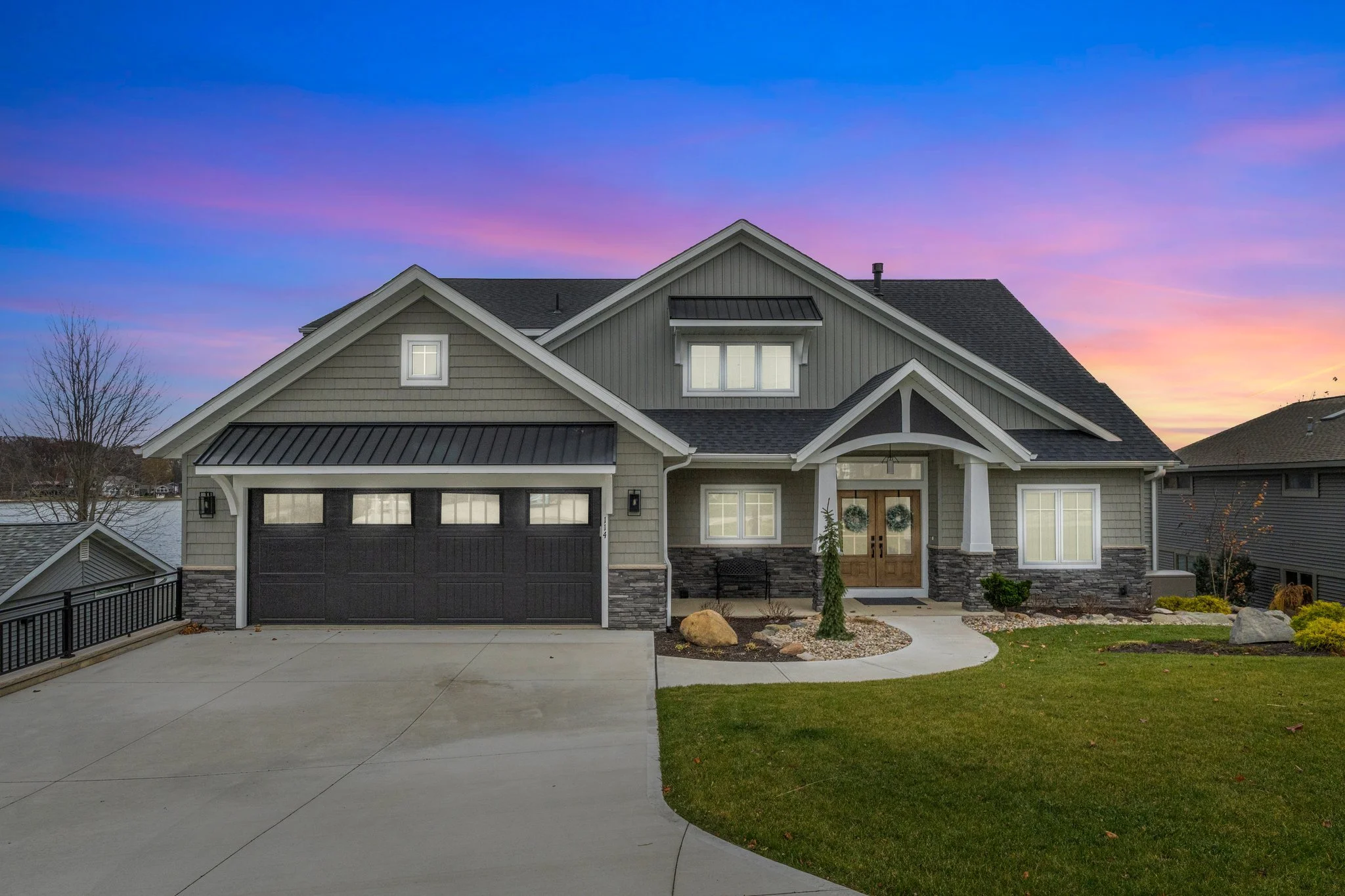 Front view of a modern suburban house with a black garage door, stone accents, and a landscaped yard during sunset.