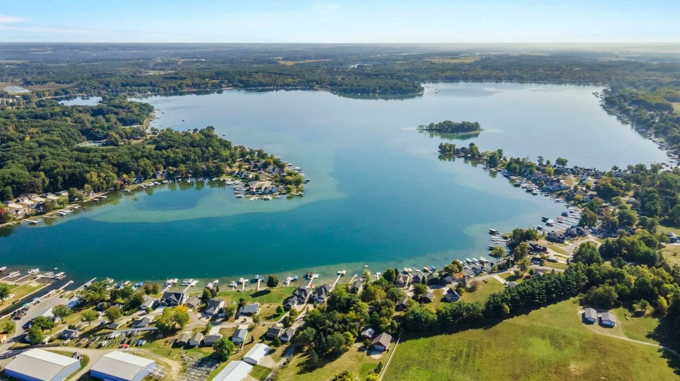 Aerial view of a large lake with houses and boats along the shoreline, surrounded by lush green trees and open fields.