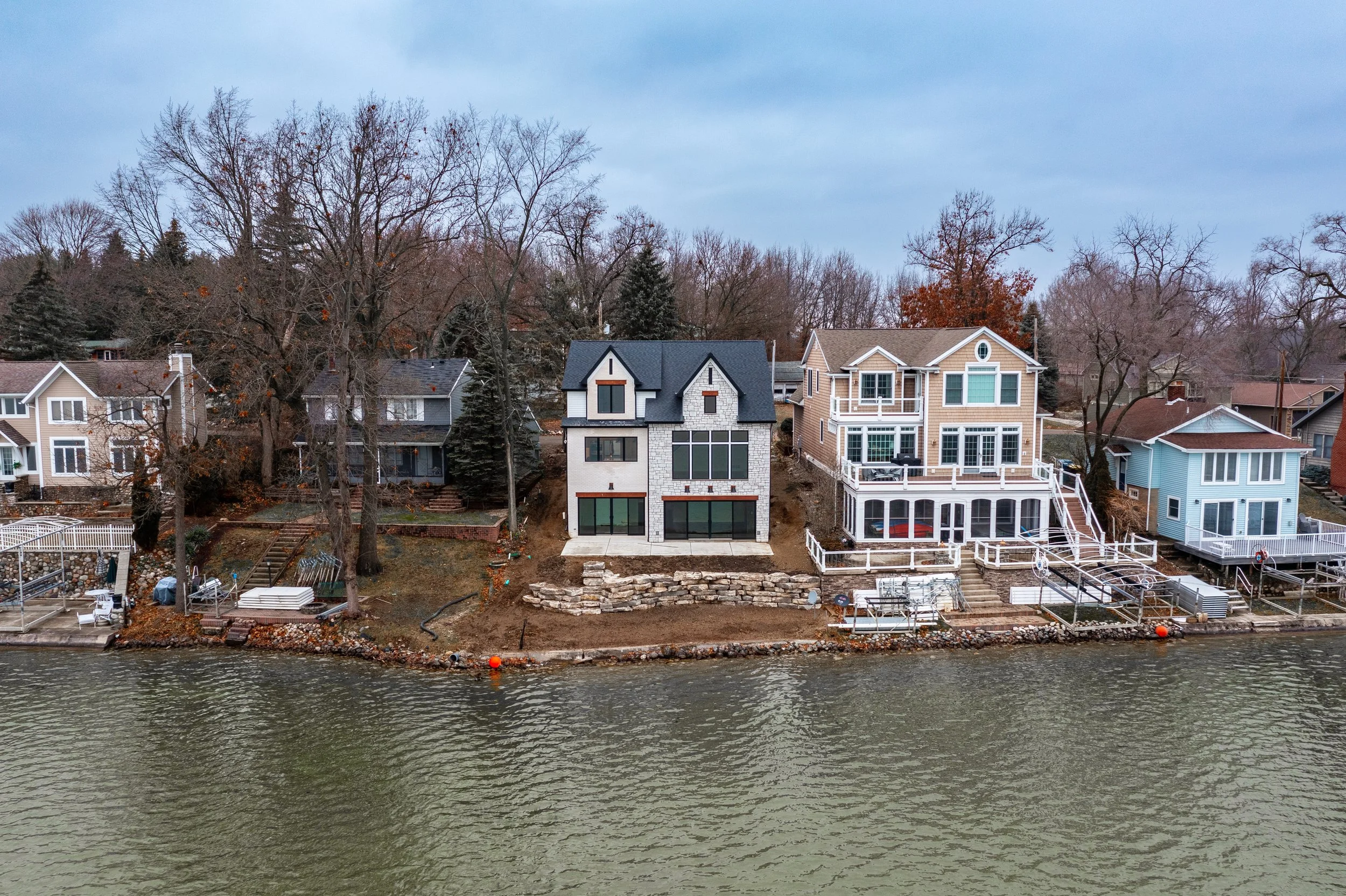 A modern house under construction on a waterfront property, with neighboring houses and leafless trees in the background.