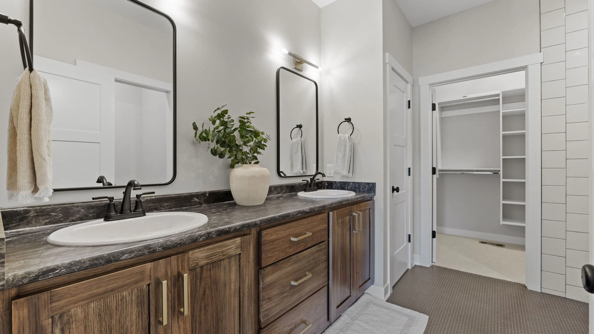 Bathroom his and hers vanity with wood cabinets, black faucets, large mirrors, a green plant in a white vase, and a walk-in closet opposite the sinks.