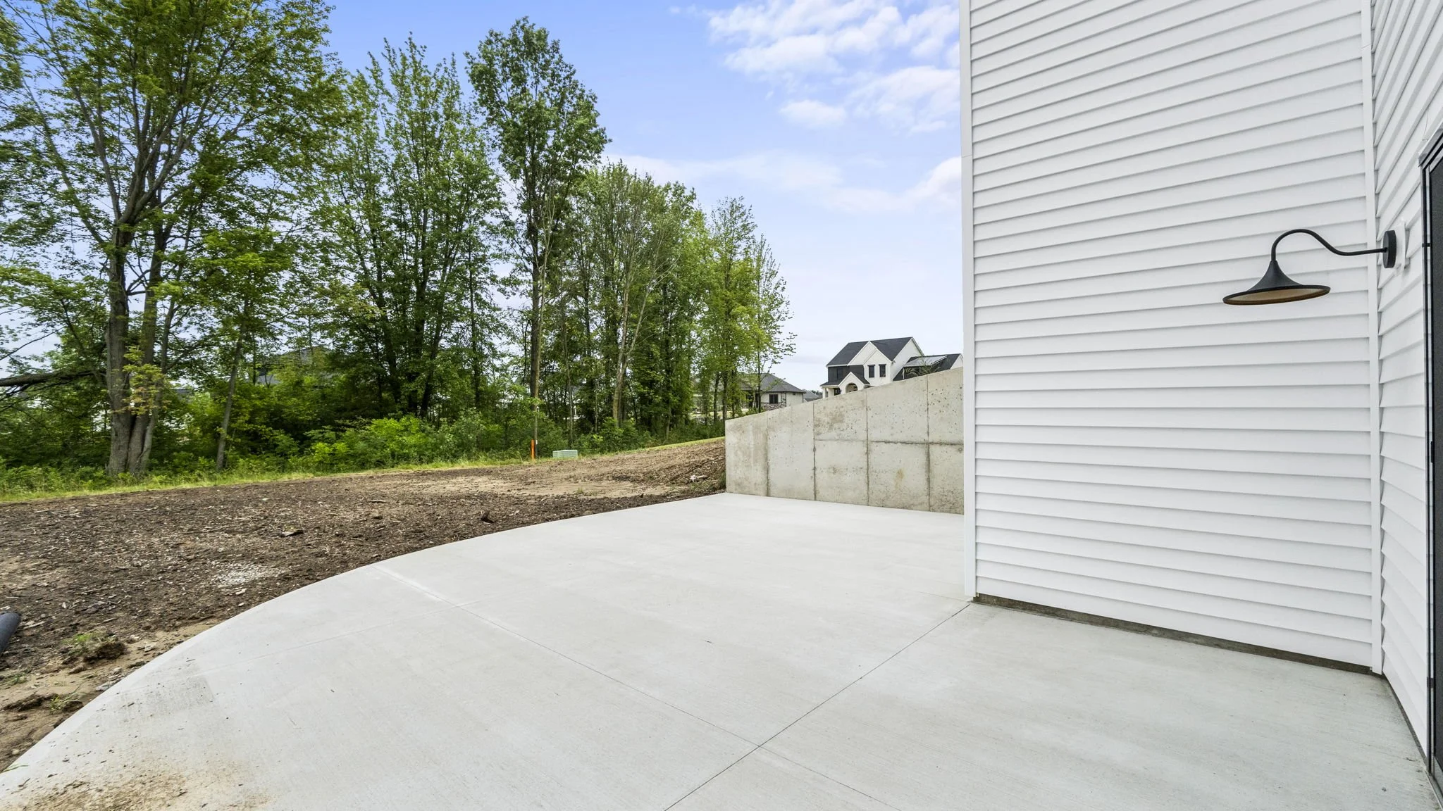 Freshly poured concrete patio with white siding house wall, outdoor light fixture, and wooded backyard in the distance.