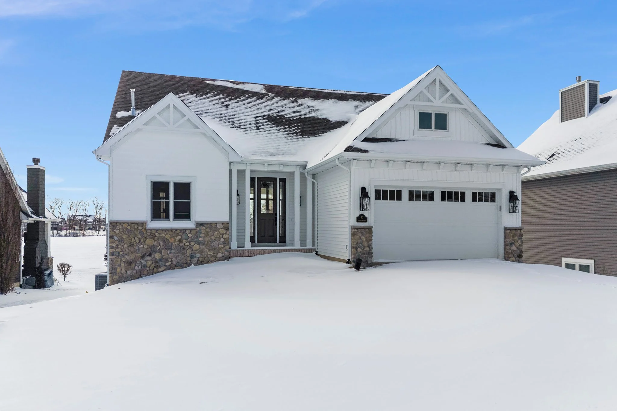 White house with stone accents on the lower walls, black front door, and garage, covered with snow in a winter setting under a blue sky.