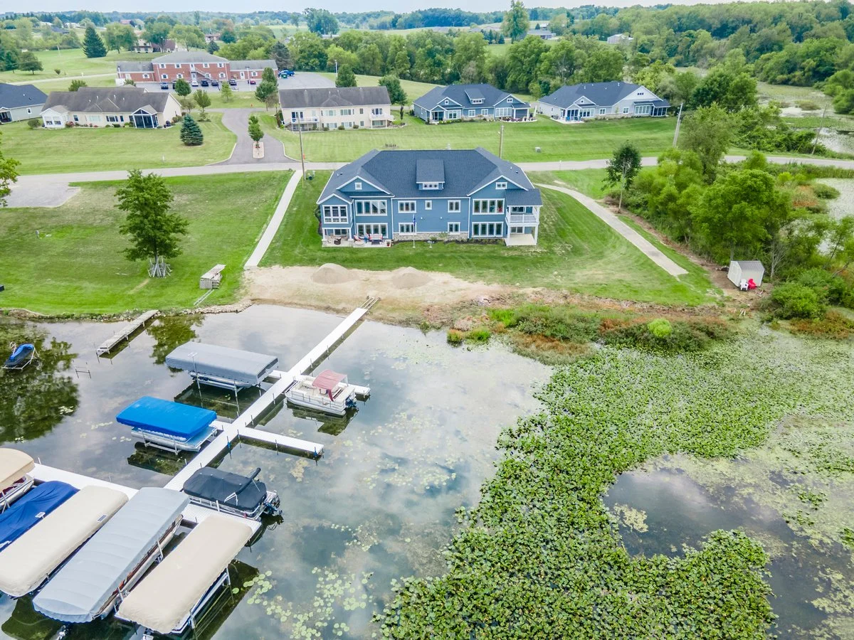 Aerial view of a lakeside residential area with a large blue house, smaller houses, boat docks, boats, lush green lawns, trees, and a waterbody with lily pads.