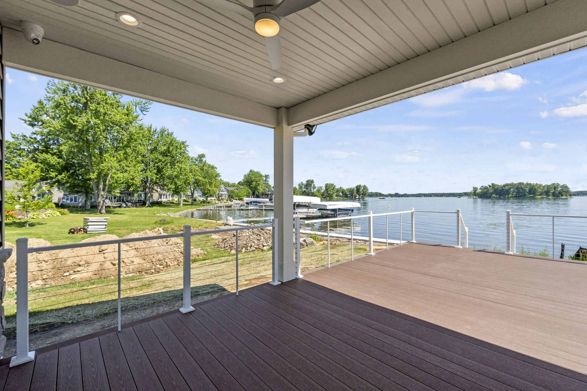View from a covered porch overlooking a lake with a dock, trees, and houses on the opposite shore, under a partly cloudy sky.