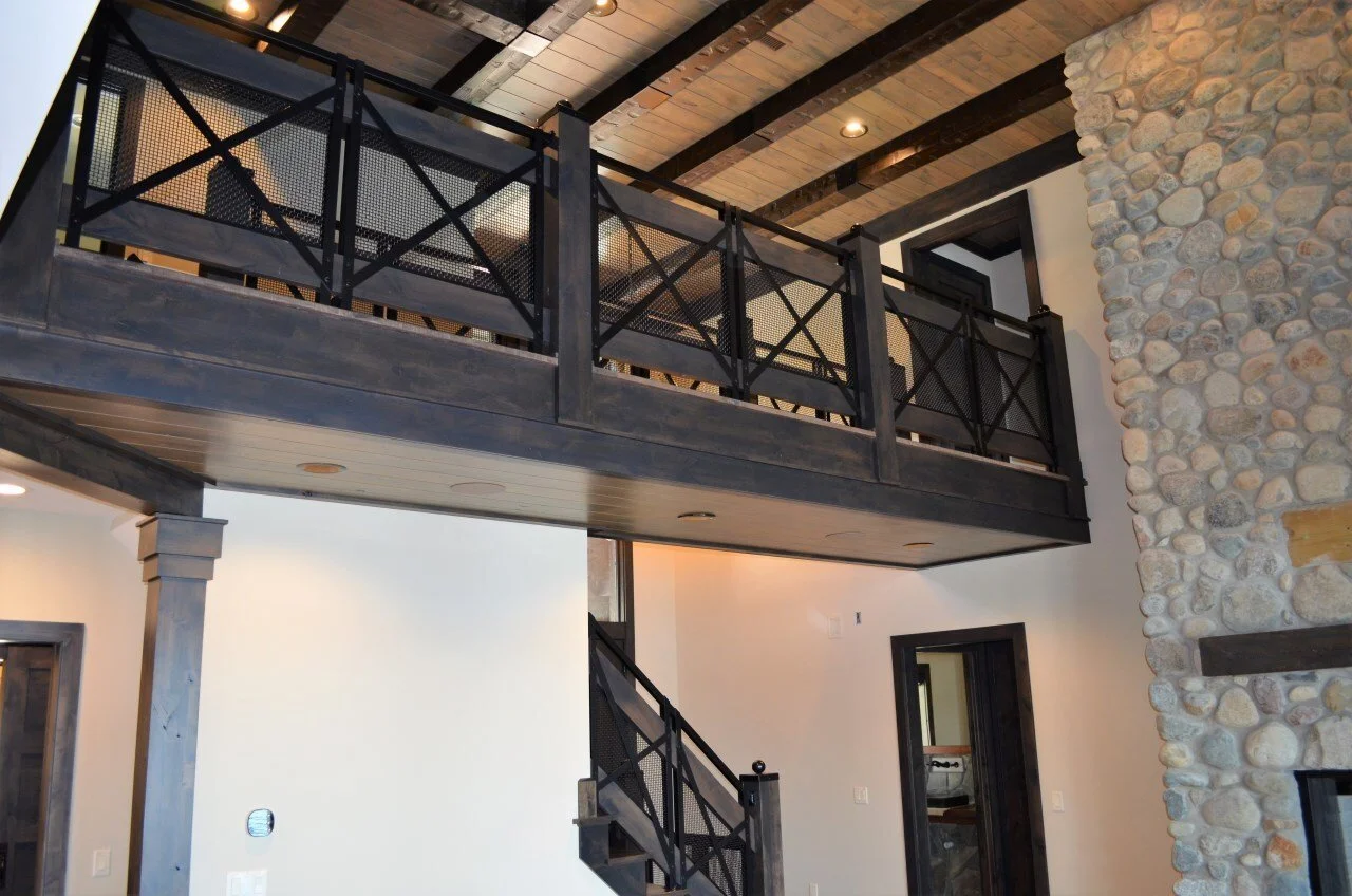 Interior of a house showing a black metal balcony railing, stone fireplace, and staircase.