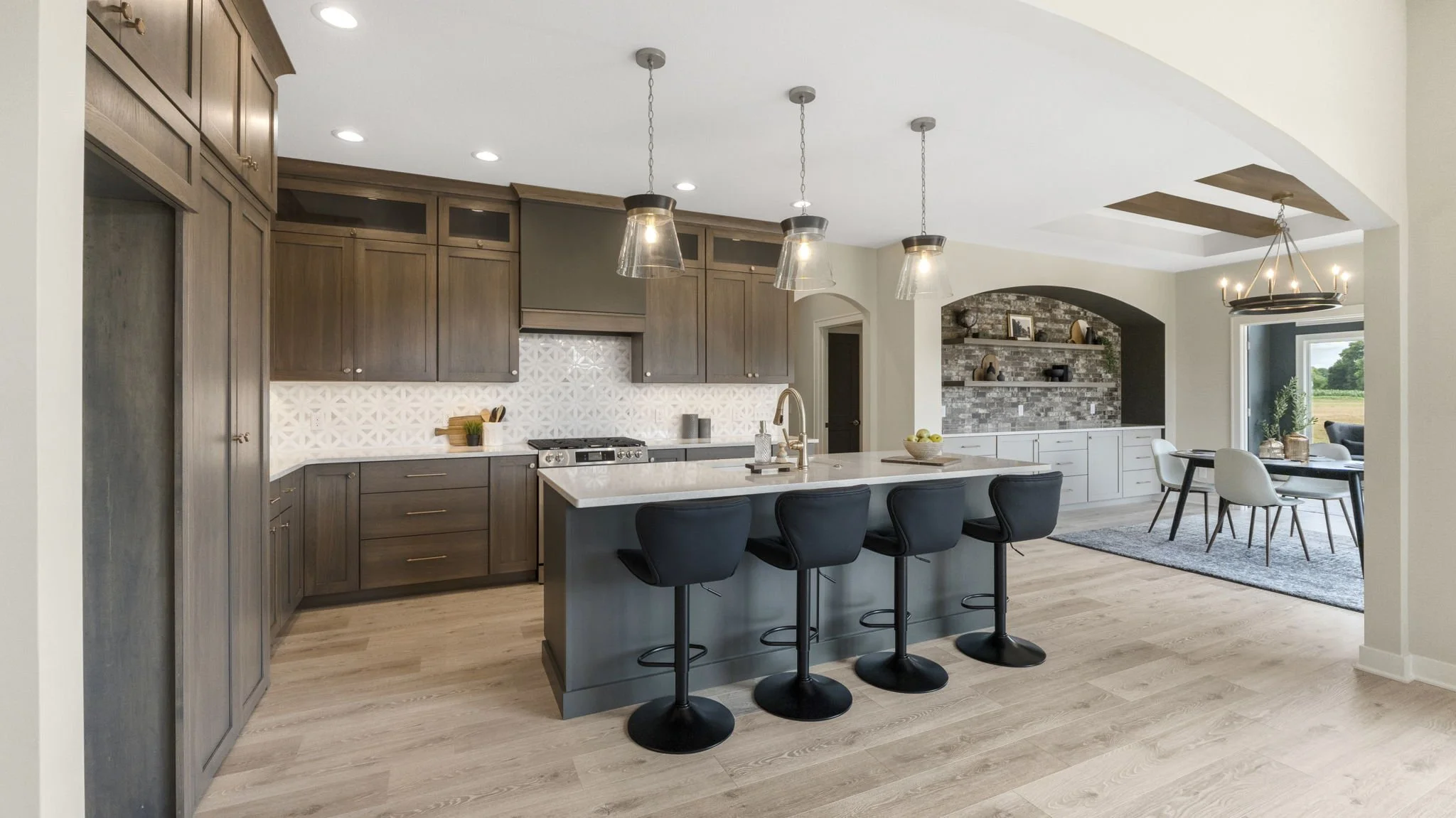 Modern kitchen with dark wood cabinets, a white tile backsplash, and a white island with four black bar stools. Adjacent dining area with a gray rug, white and black chairs, and a brick accent wall with built-in shelves.