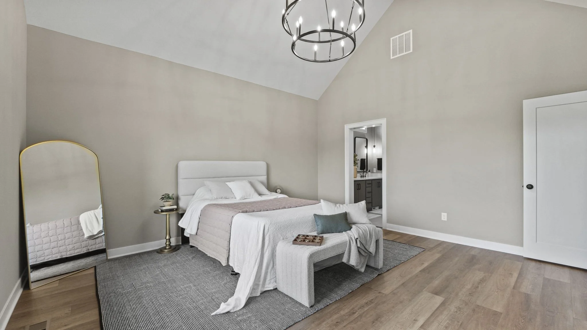 A bedroom with a white bed, gray and pink bedding, a bench at the foot of the bed, a mirror leaning against the wall, and a doorway leading to a bathroom with black cabinets and a sink.