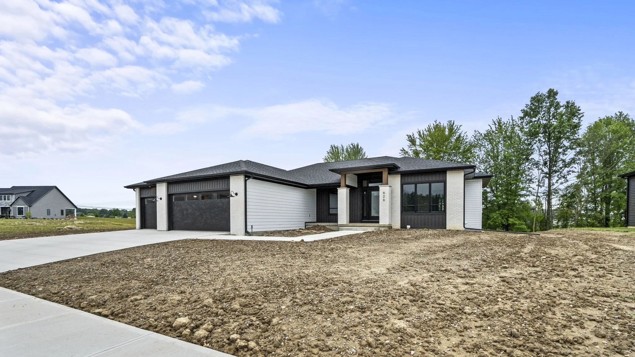 Newly built modern single-story house with black and white exterior, black garage doors, and a small front porch with steps, surrounded by dirt yard and some trees in the background.