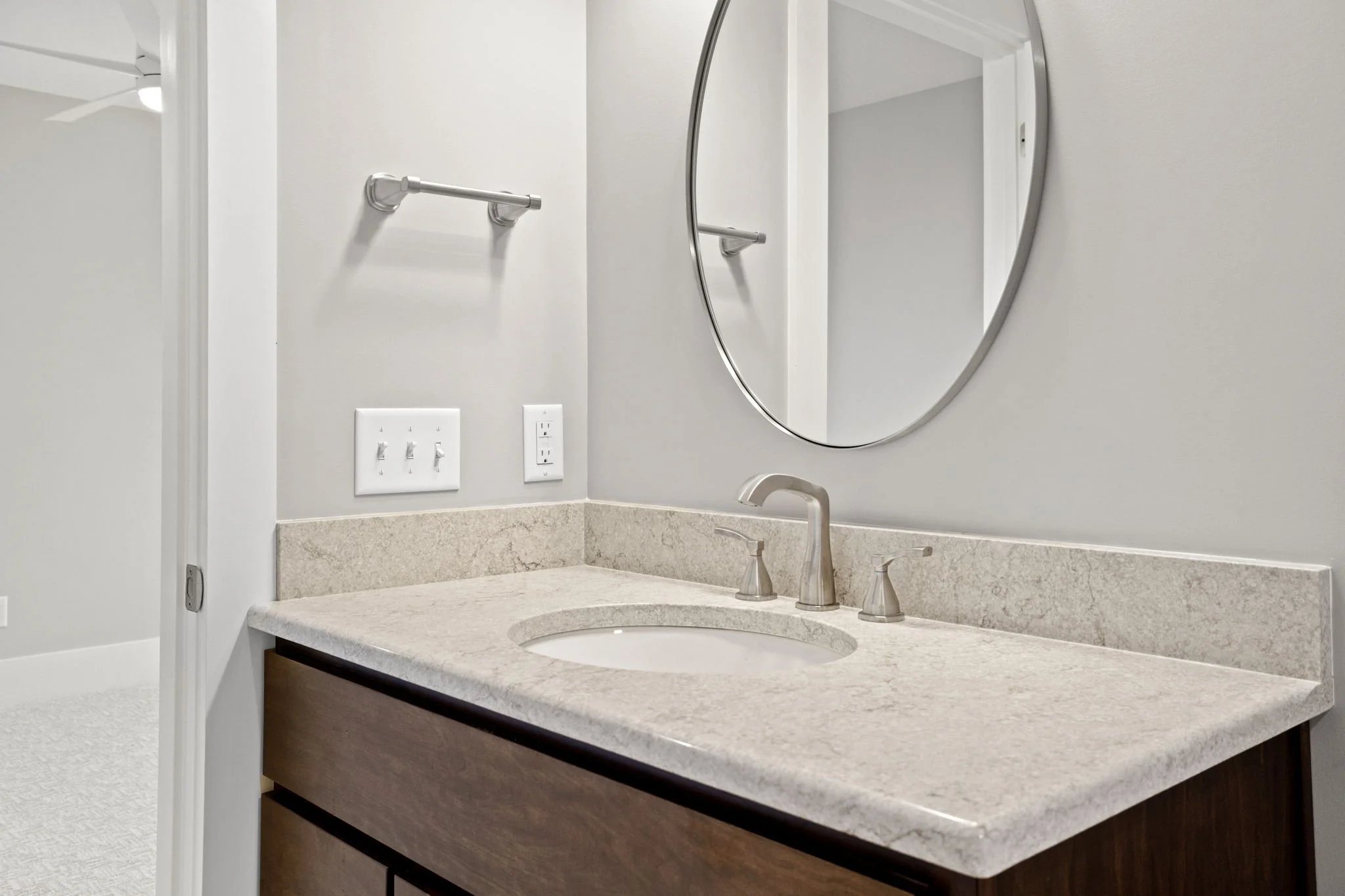 Bathroom vanity with a round mirror, a beige granite countertop, and a faucet. There is a towel bar, electrical outlets, and light switches on the wall.