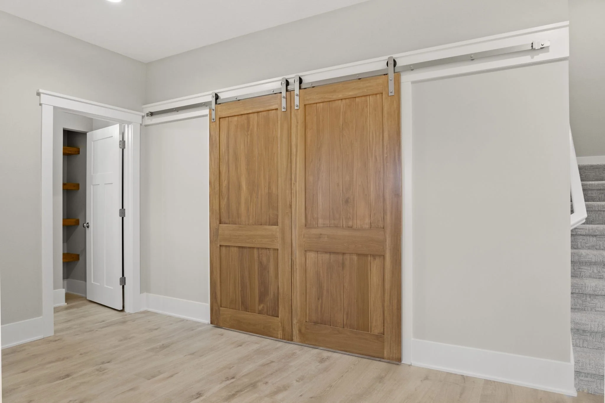 Interior view of a room with white walls, a light wooden floor, a door, and a sliding wooden barn door on a metal track.