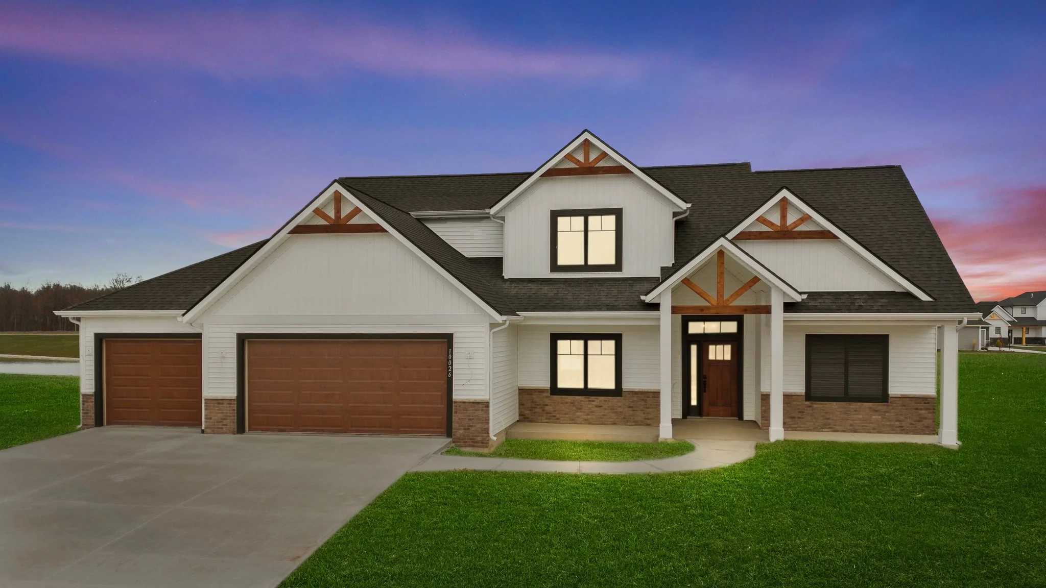 Modern two-story house with white siding, brick accents, and wooden garage doors, set against a colorful sunset sky with green lawn.