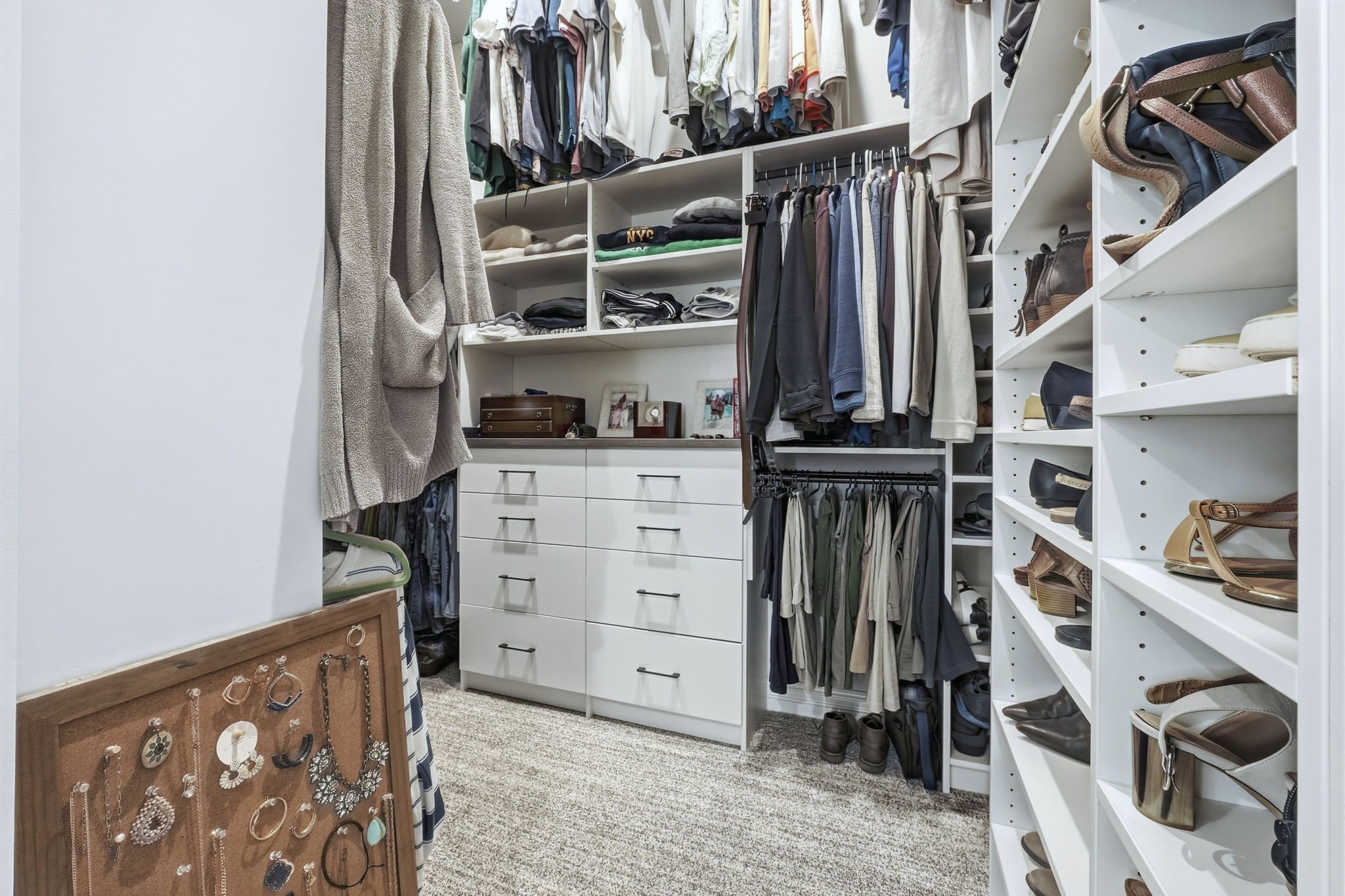 A walk-in closet with neatly organized shelves of clothes, shoes, and accessories. There are folded shirts, hanging pants, and a collection of shoes on the right shelves. A jewelry display board with earrings and necklaces is on the left side.
