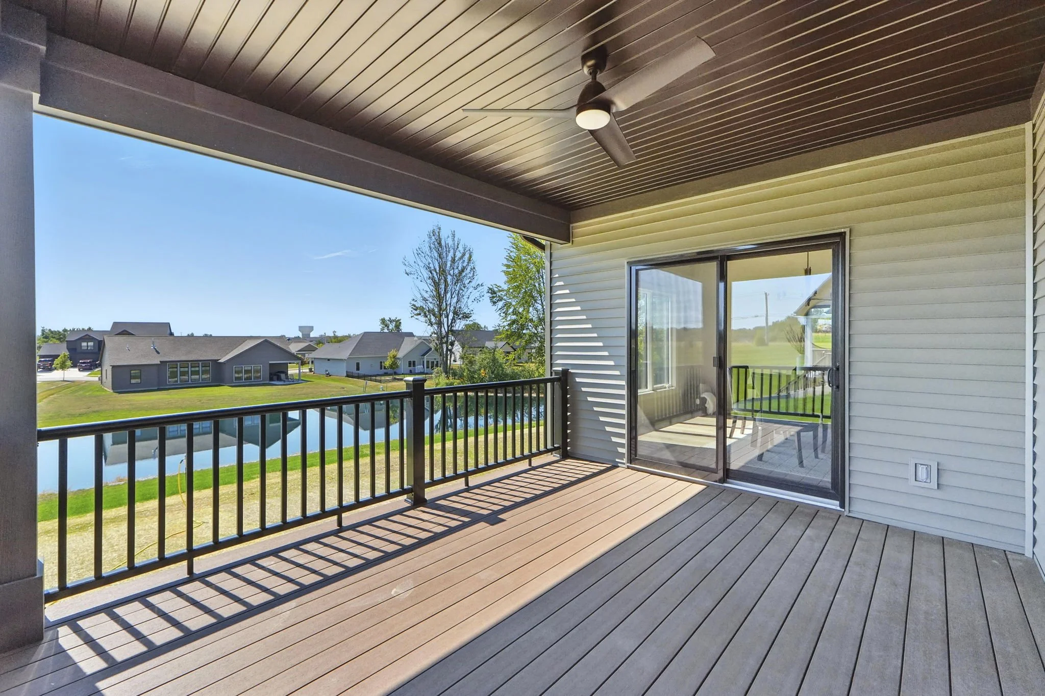 View of a porch with wood decking, black railing, ceiling fan, and sliding glass door leading to the interior. Overlooks a pond and neighboring houses under a clear blue sky.