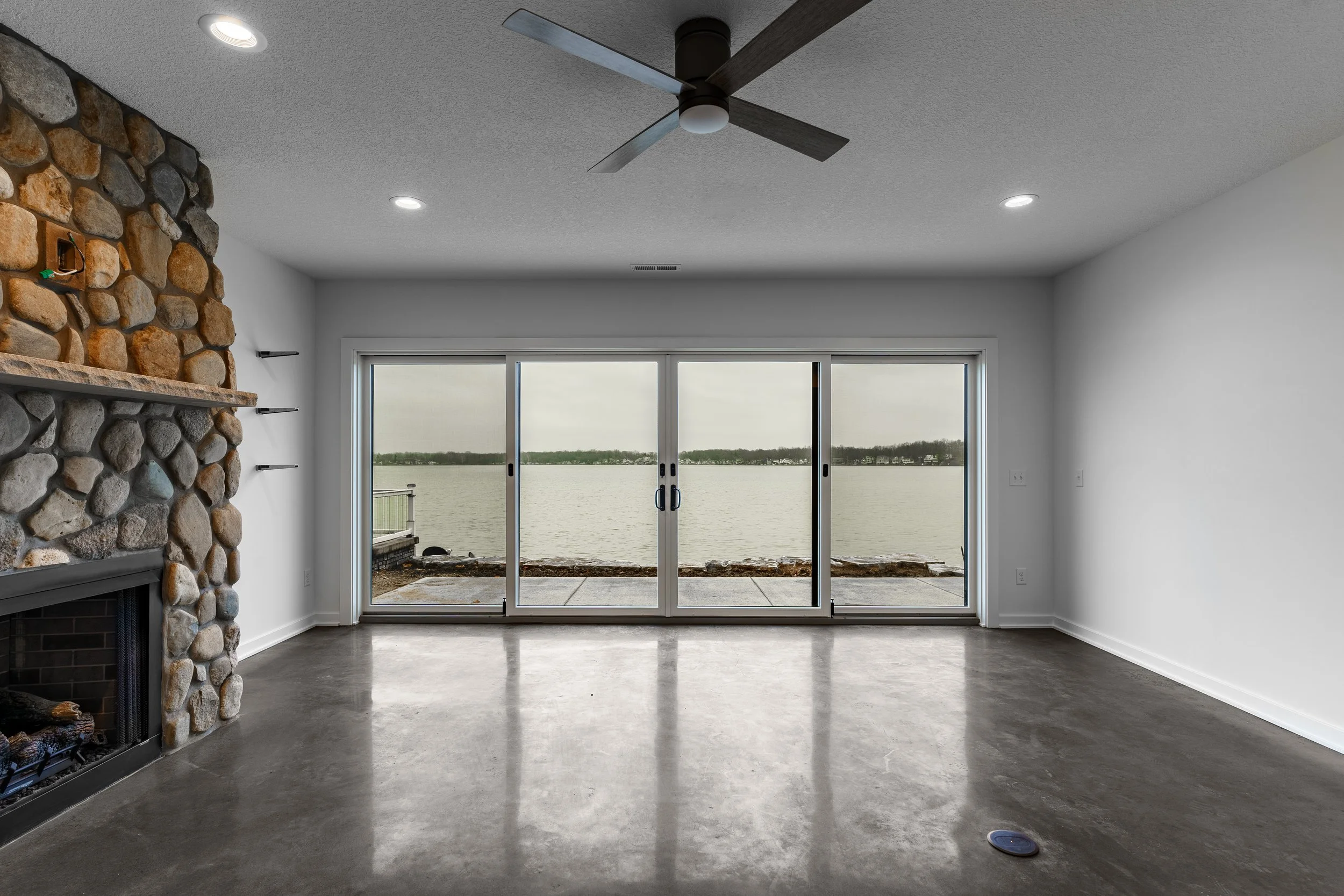 Empty living room with a stone fireplace on the left, large glass sliding doors in the center, and a ceiling fan overhead, overlooking a body of water outside.