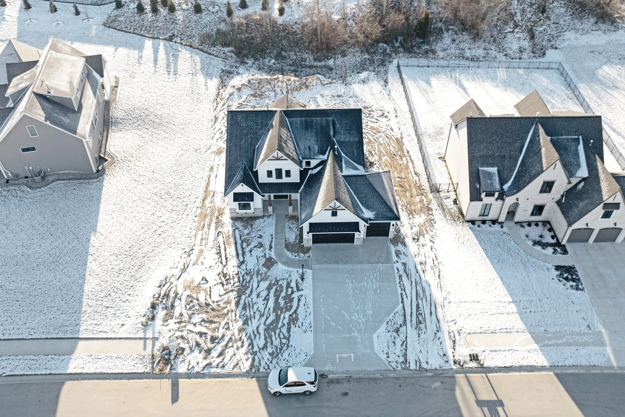 An aerial view of a suburban neighborhood showing snow-covered lawns and rooftops. The central house is a large, modern two-story home with a driveway and a parked car in front.