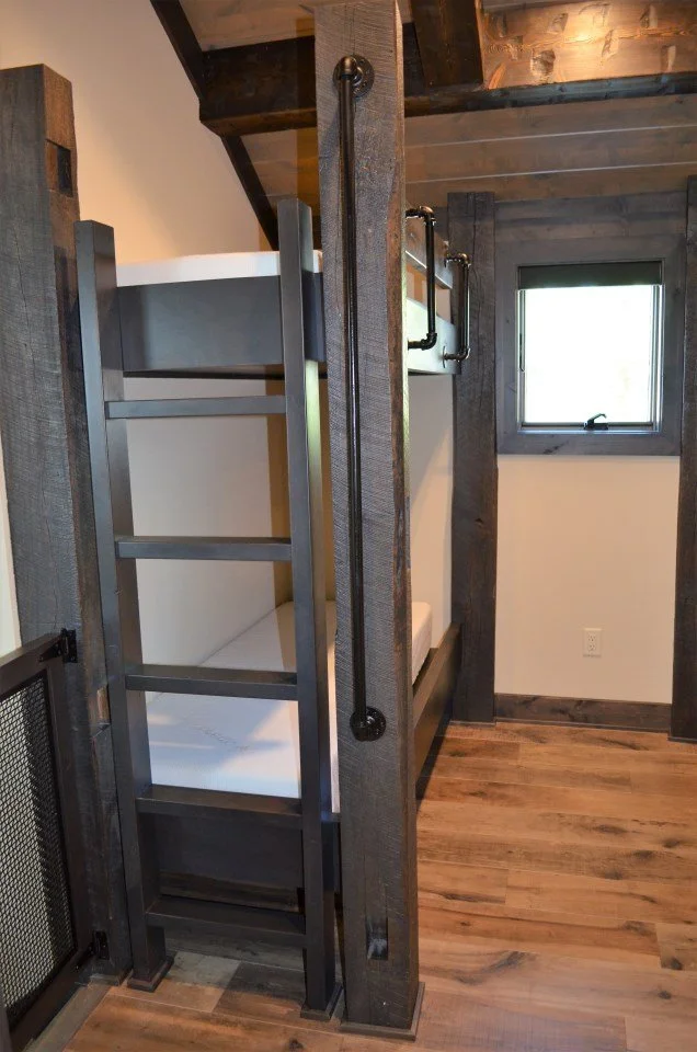 A small bedroom with wooden floors and walls, featuring a bunk bed next to a window, with a black metal ladder and safety rail.