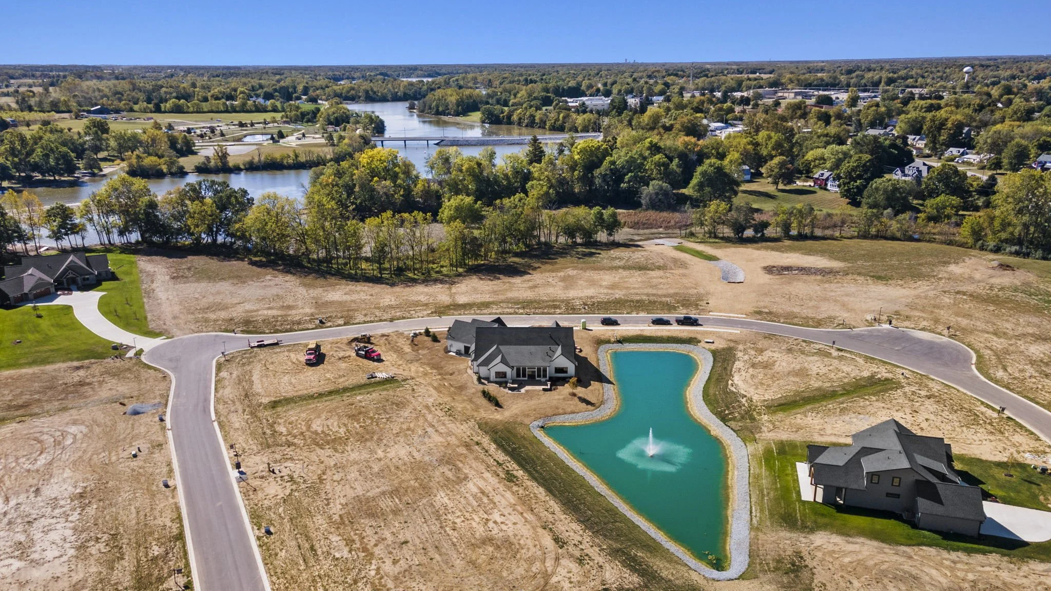 An aerial view of a suburban area with a house, a pond with a fountain, roads, and undeveloped land. There is a river in the background surrounded by trees.