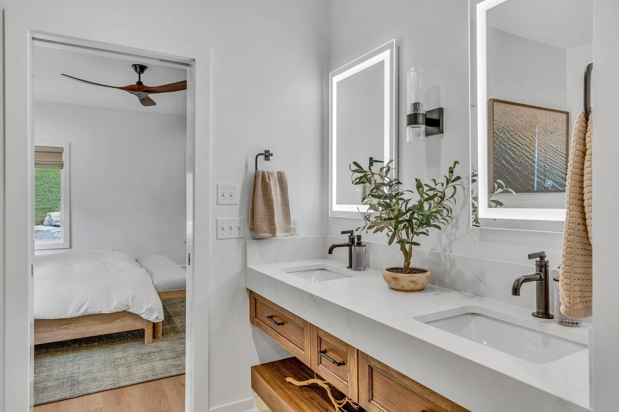 Modern bathroom with double sinks, illuminated mirrors, a potted plant, and beige towels, adjacent to a bedroom with a bed, window, and ceiling fan.