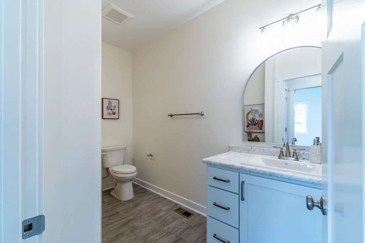 A small bathroom with a white toilet, a white vanity with a marble top, a large mirror, a wall art picture, a towel bar, a soap dispenser, a vent on the ceiling, and a window with sunlight