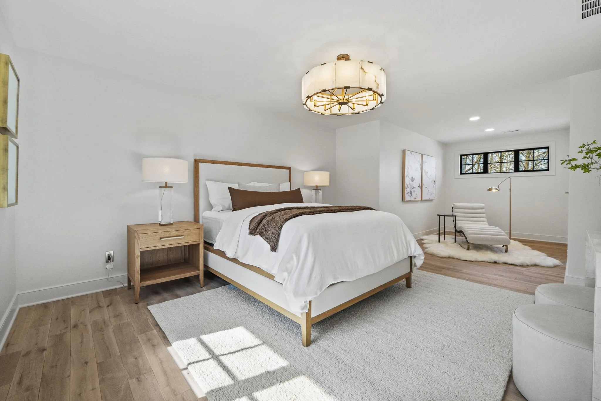 Bright modern bedroom with white walls, wooden floors, a wooden bed frame with white bedding, brown throw blanket, and pillows. Nightstand with a lamp on each side of the bed, ceiling light fixture, a cozy area with a lounge chair, small table, floor