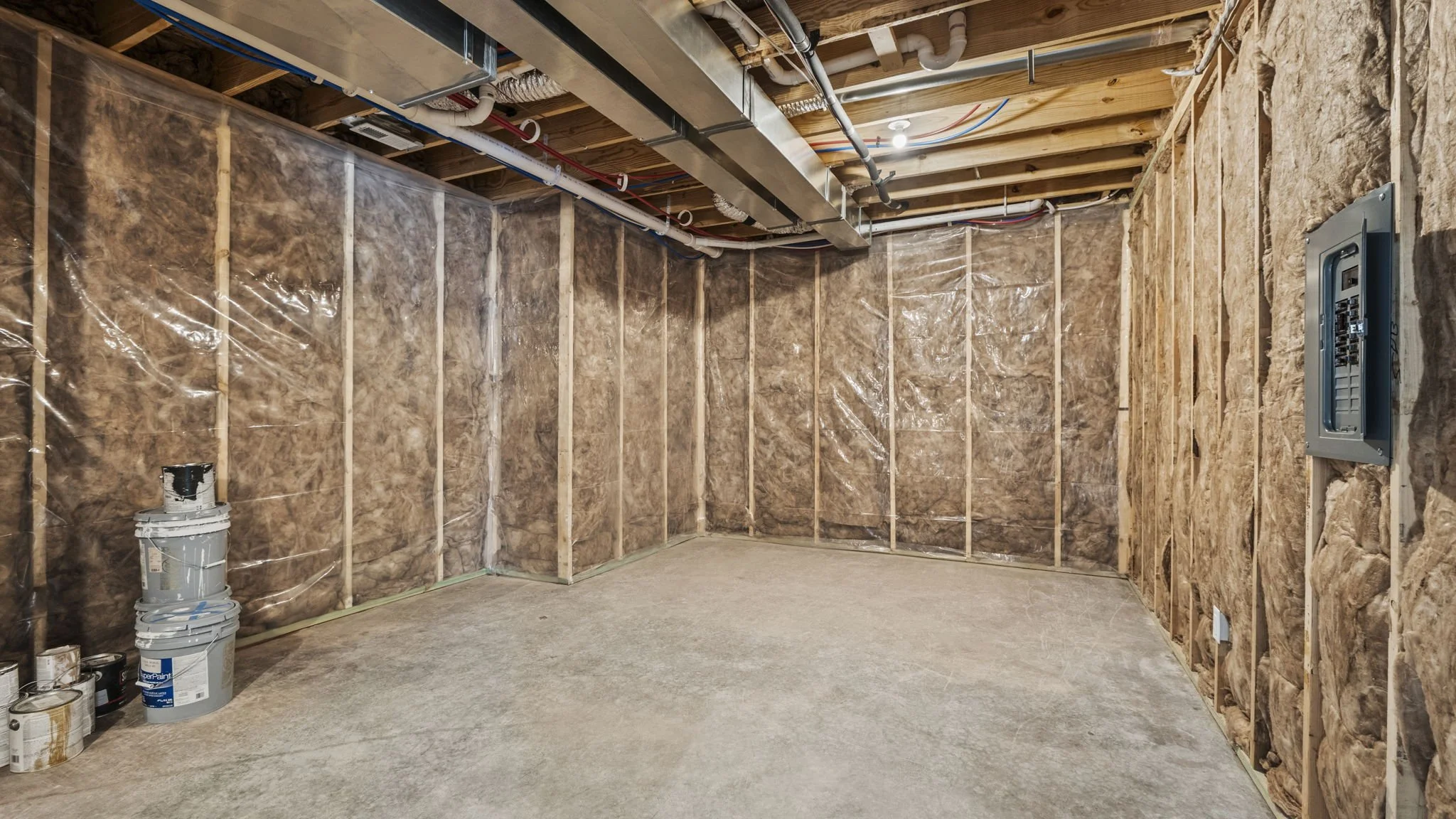 An unfinished basement with insulation installed on the walls, exposed ceiling with visible pipes and ducts, an open electrical panel, and some paint cans and supplies on the floor.