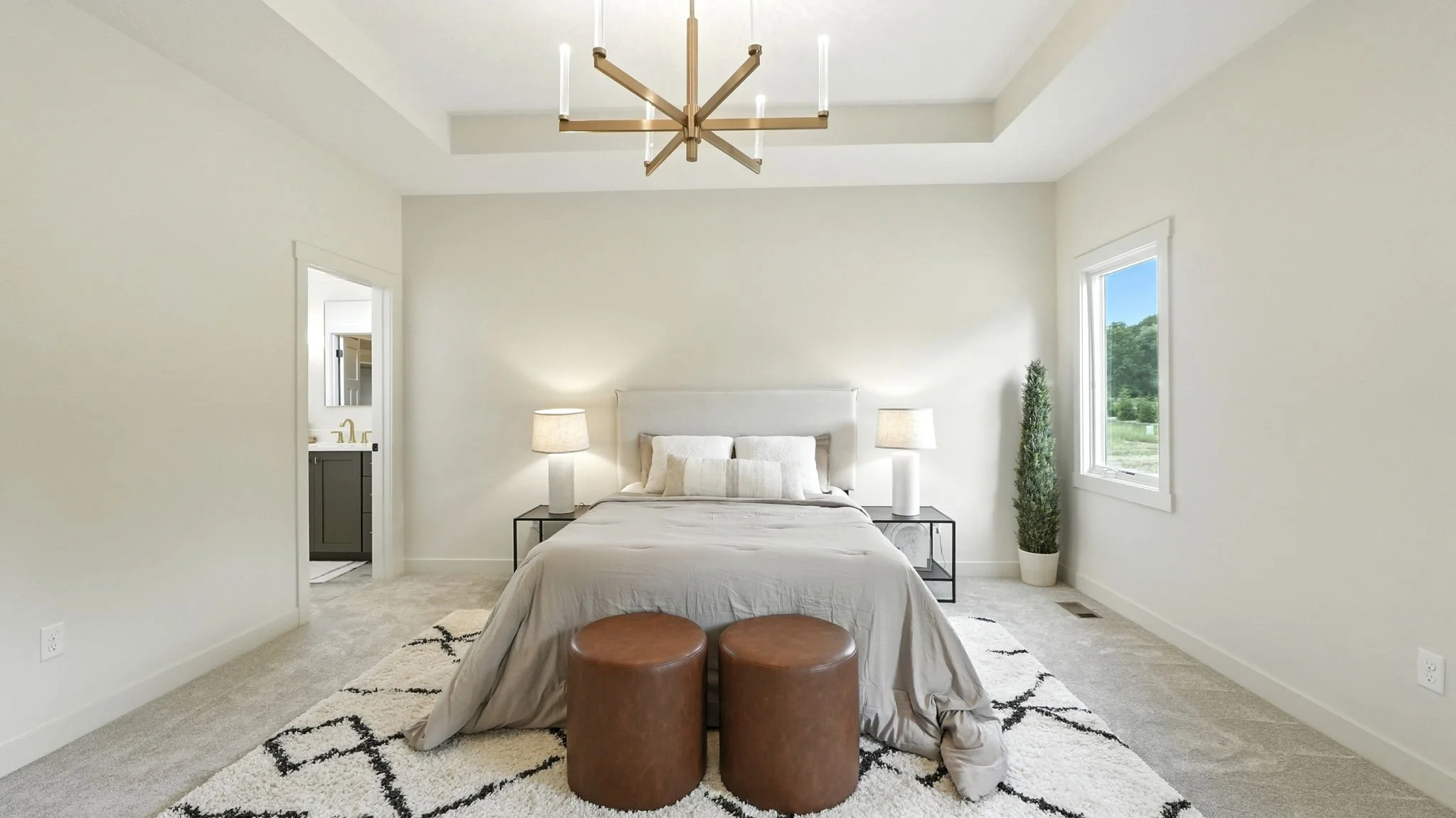 A modern bedroom with white walls, a bed with white bedding and pillows, two bedside lamps, a black metal nightstand, a tall green plant in a white pot, a window showing greenery outside, leather ottomans at the foot of the bed, and a geometric area 