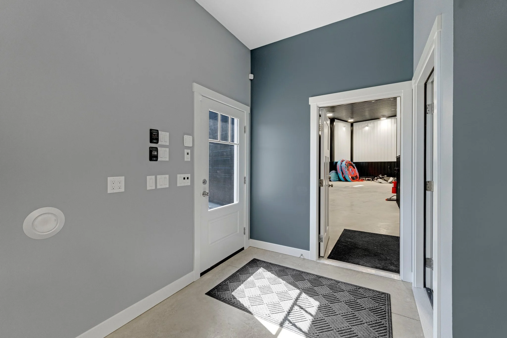 Entryway of a house with a gray wall, a white door, and an open doorway leading to a storage or utility area with sports equipment.