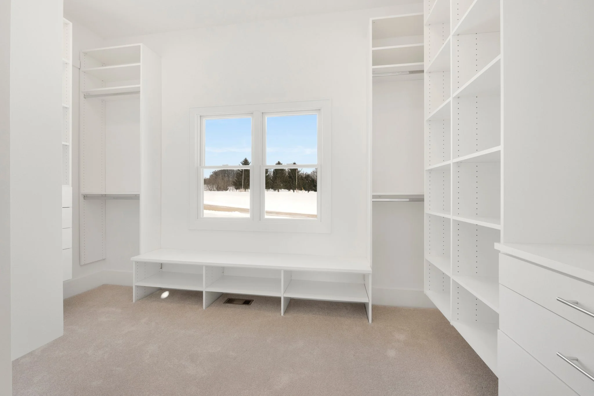 Empty walk-in closet with white built-in shelves and drawers, beige carpet, and a window showing an outdoor snowy landscape.
