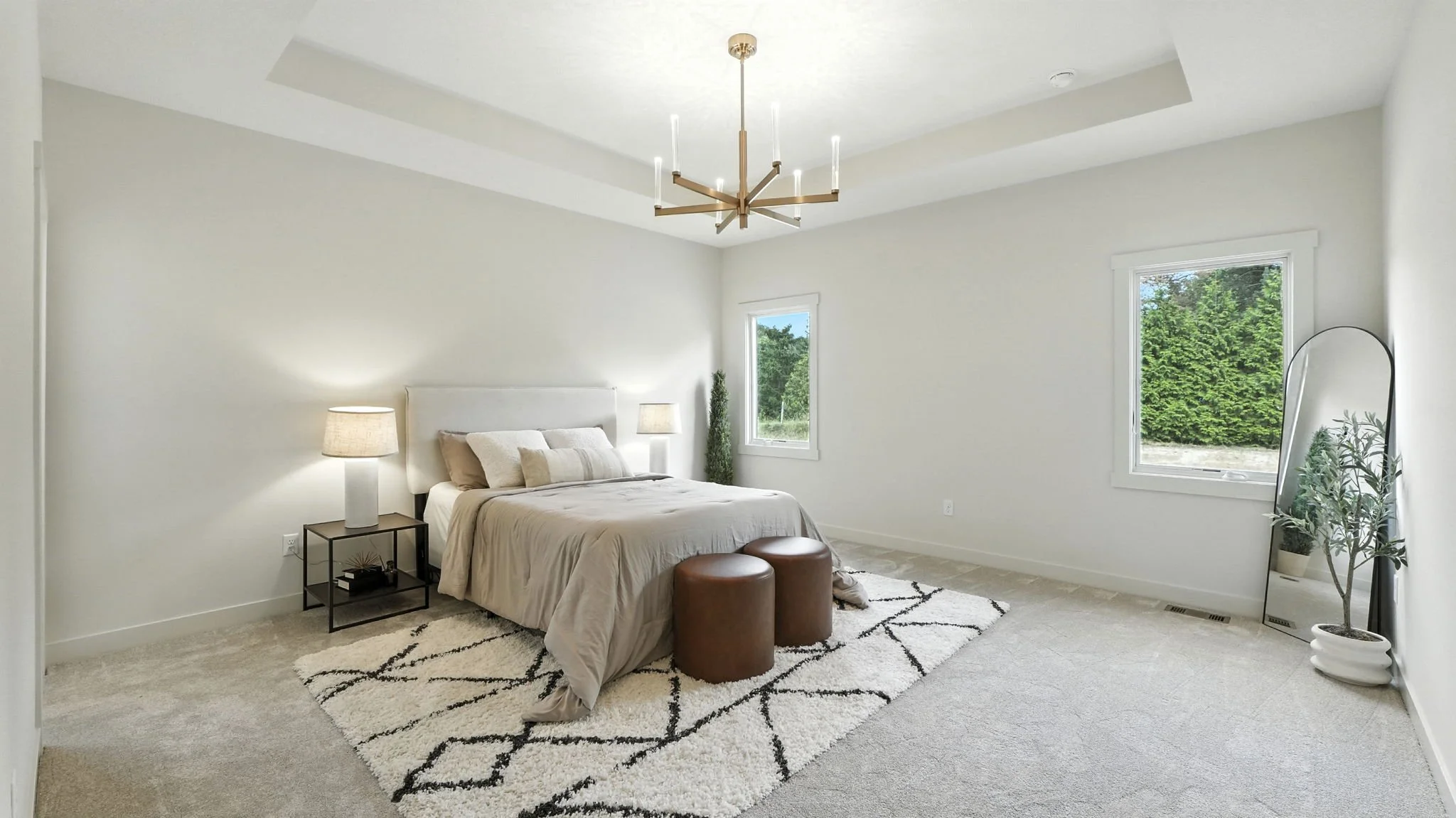 A minimalist bedroom with white walls, beige bedding, two brown ottomans at the foot of the bed, a black nightstand with a lamp, a large mirror, and two windows showing greenery outside.