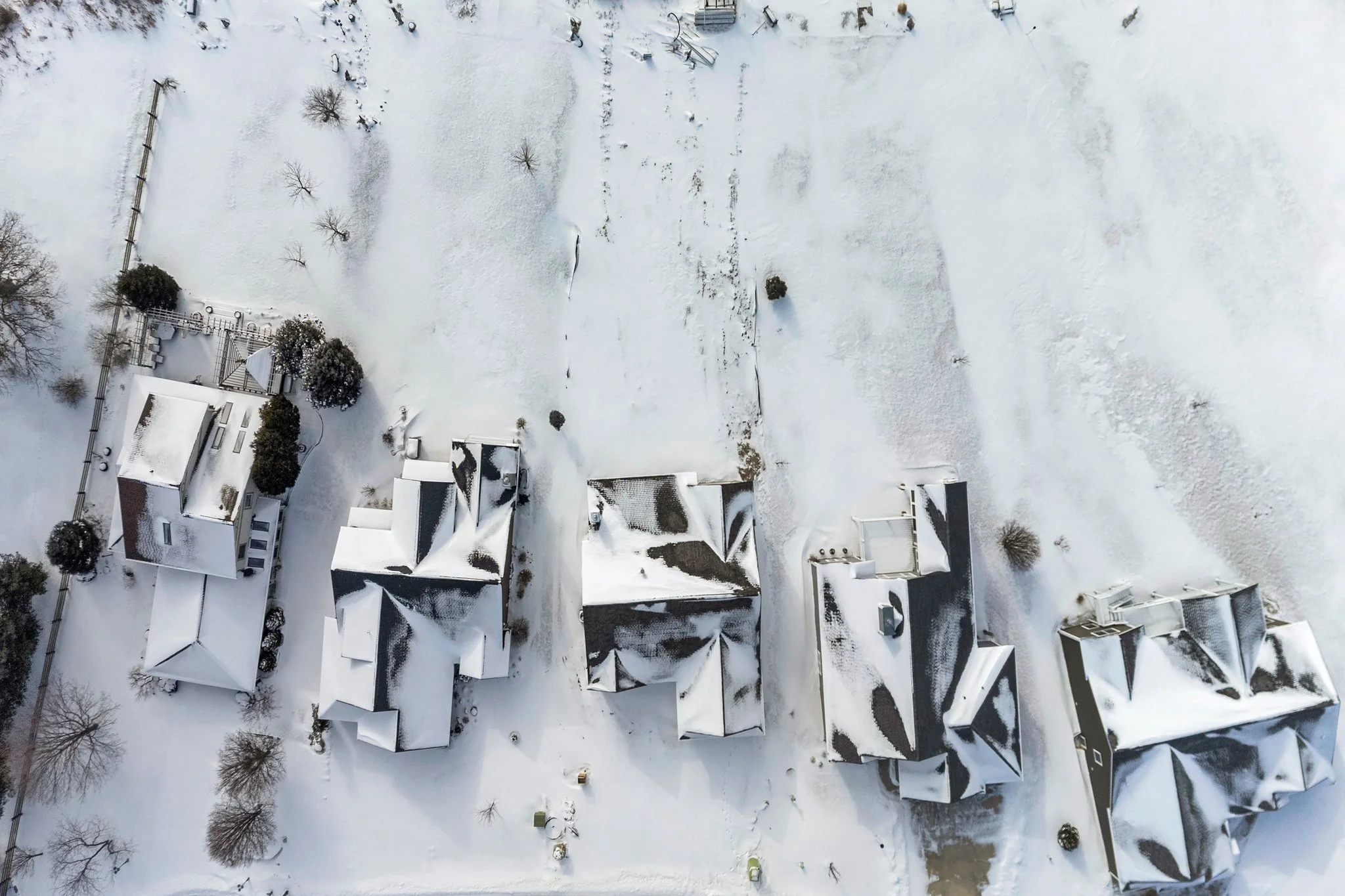 An aerial view of five houses with snow-covered roofs in a residential area, trees, and a snow-covered lawn.