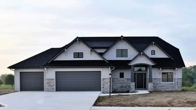 Modern two-story house with a black roof, white and stone exterior, front door, three garage doors, and some landscaping.