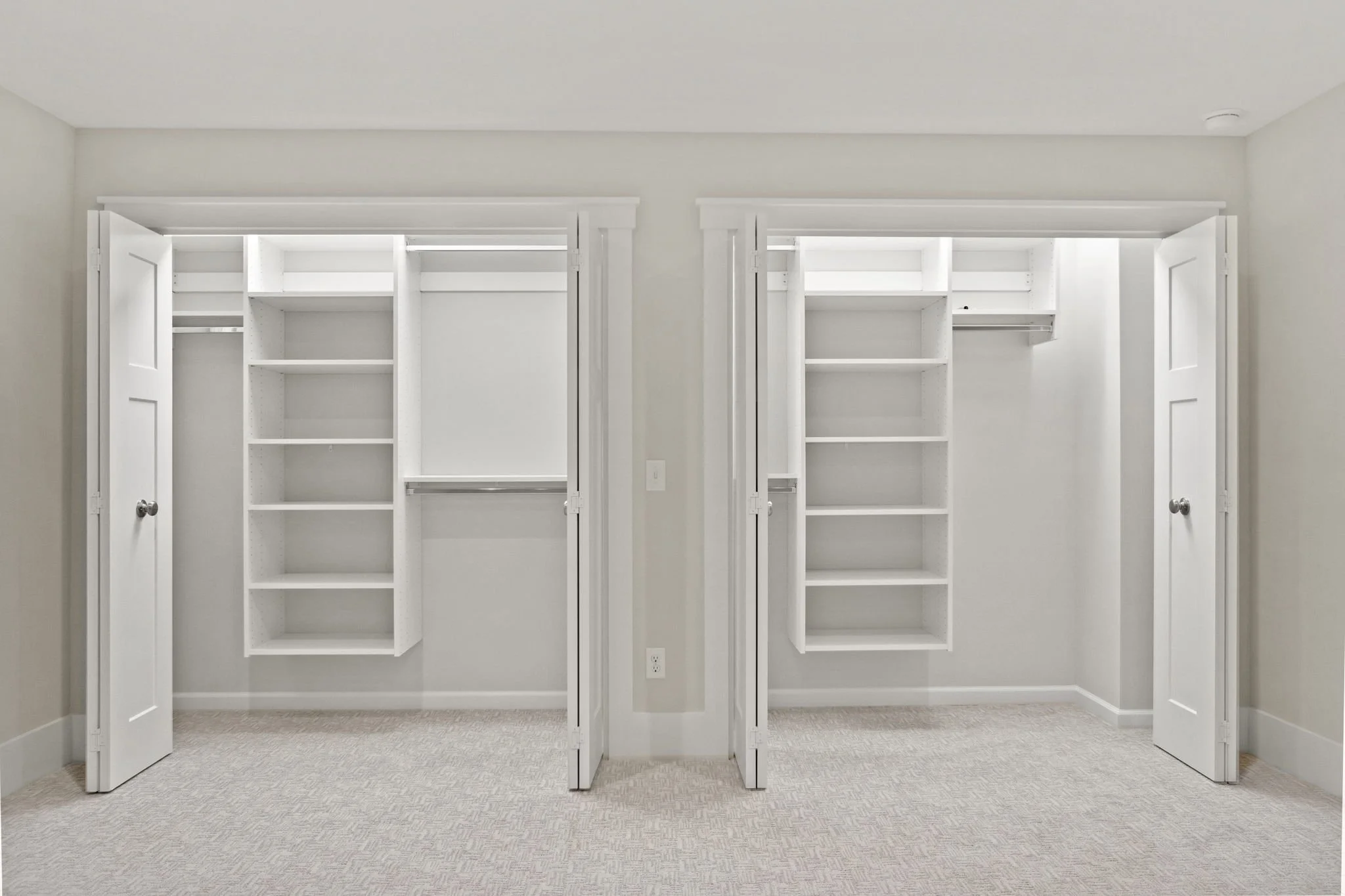 Empty walk-in closets with white shelving and hanging rods, open doors, beige carpet, and plain white walls.