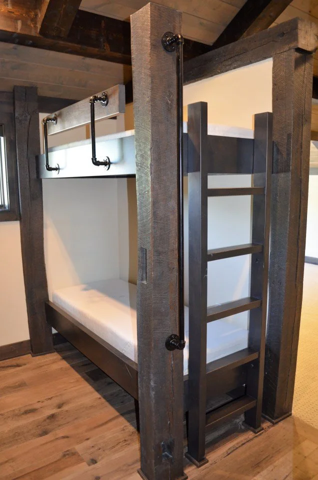 A bunk bed made of wood with a rustic finish, featuring a ladder on the right side and a safety rail on the upper bunk, located in a room with wooden flooring and a ceiling with exposed beams.