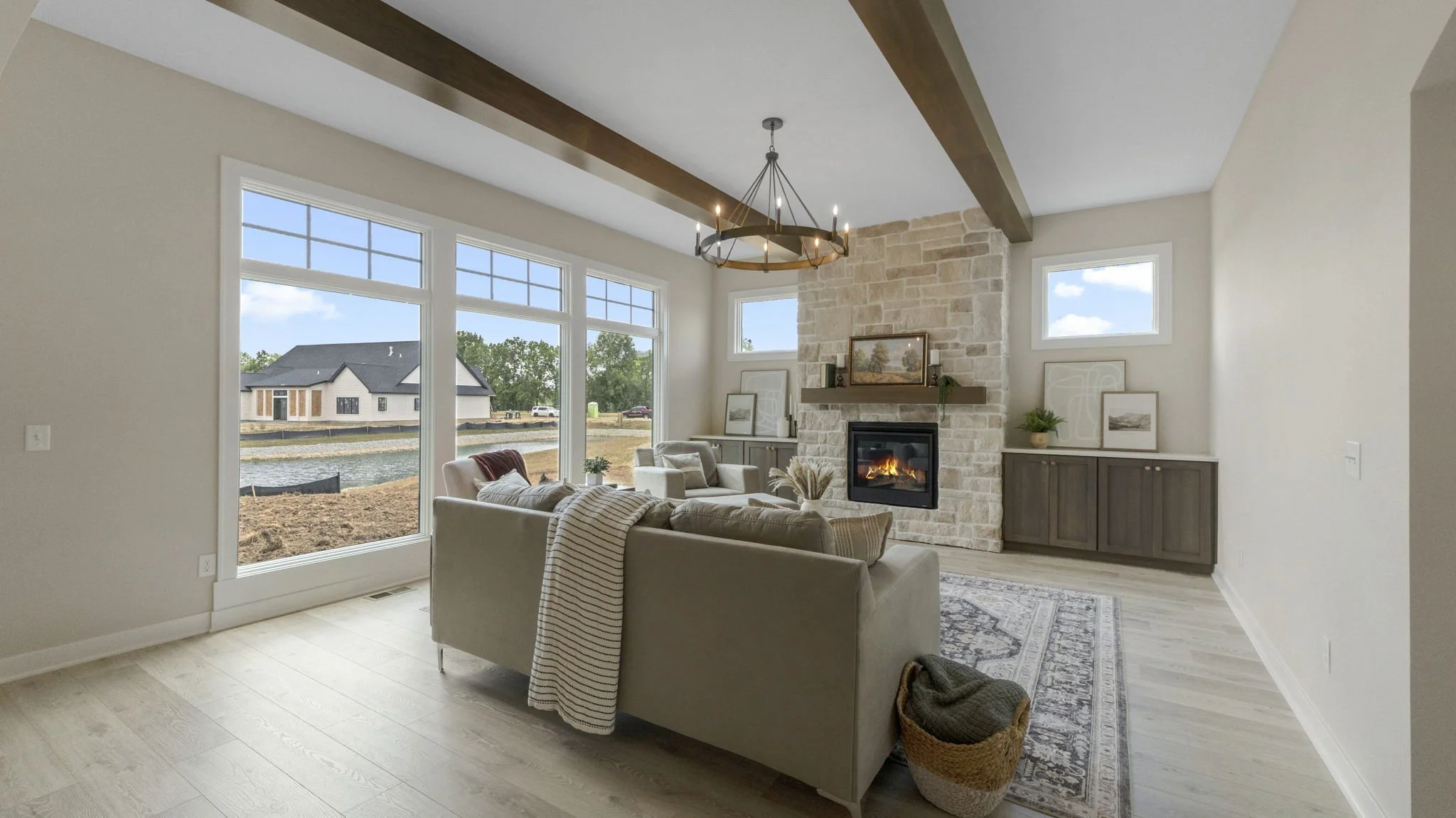Living room with beige couch, white and beige armchairs, stone fireplace, large windows showing yard and pond, wooden ceiling beam, chandelier, and decorative art pieces.