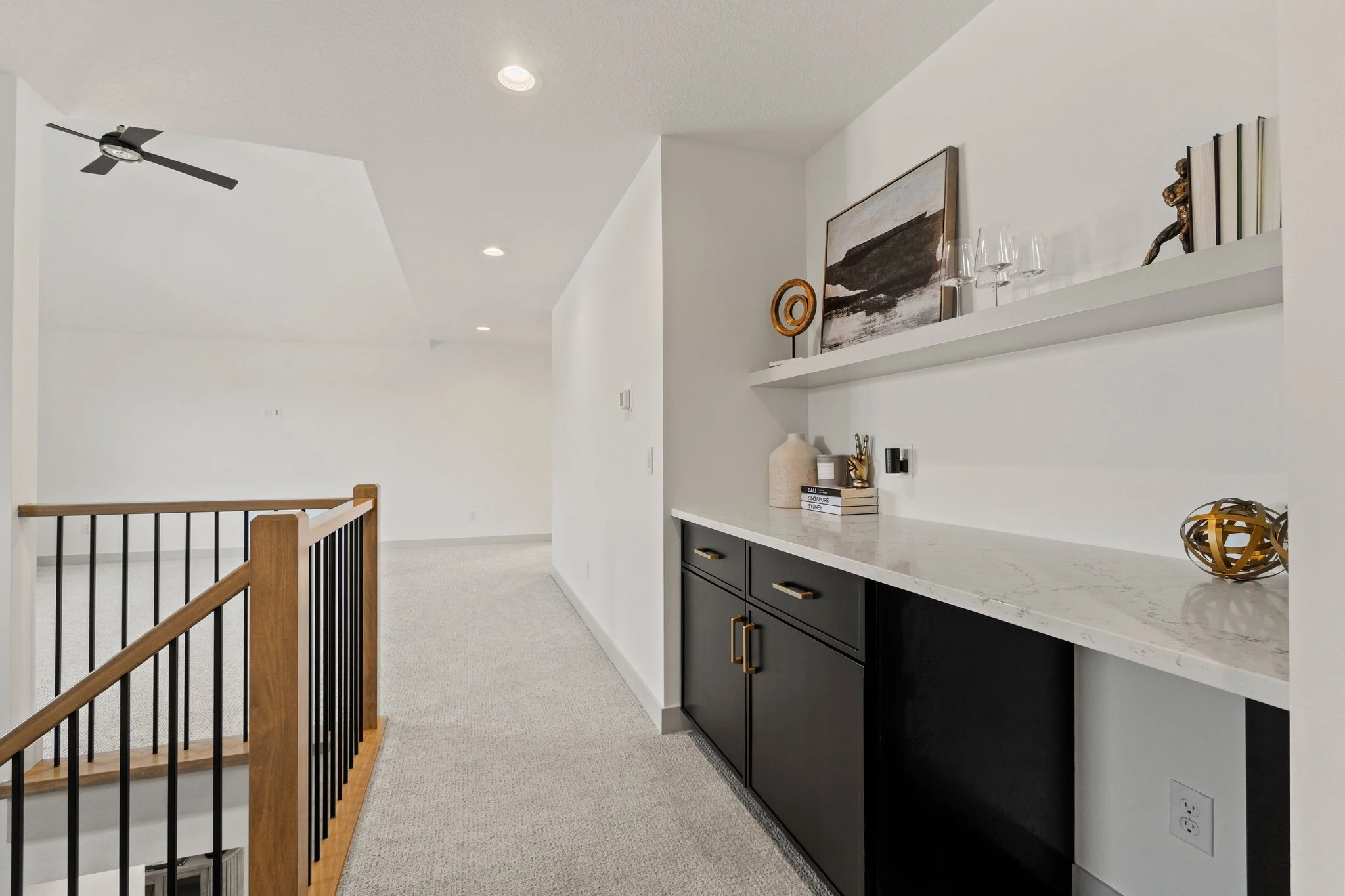 Interior view of a modern home with a staircase, a white wall with a black cabinet, decorative items, books, artwork, and glassware on a floating shelf, and ceiling lights.