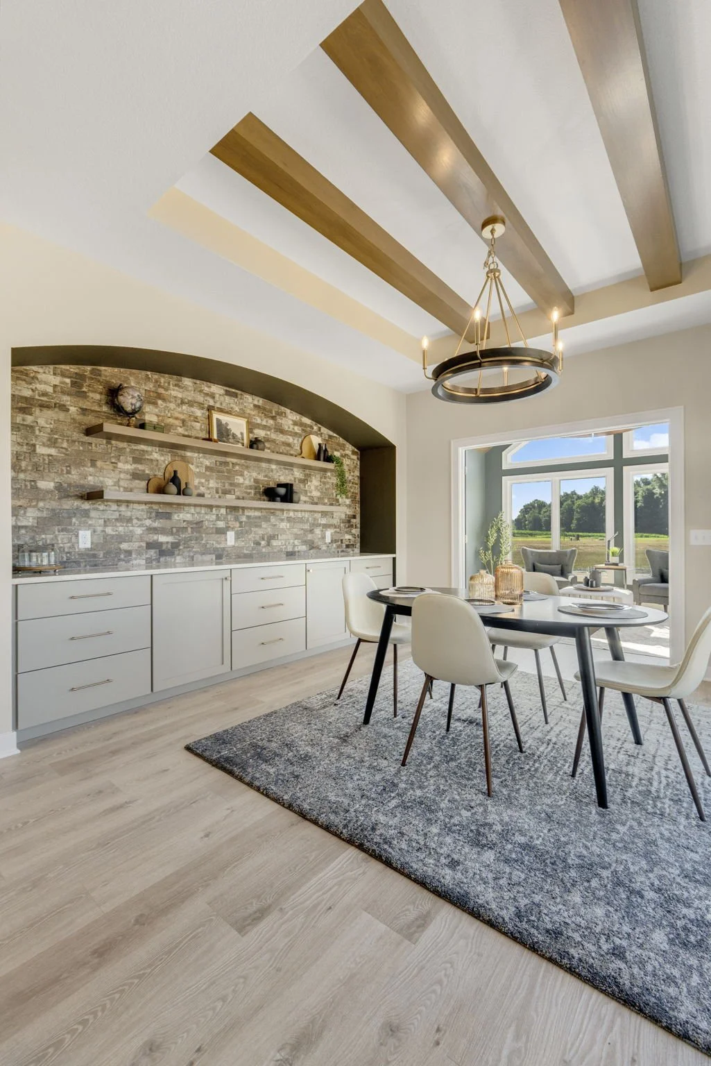 Modern dining room with a black round table, cream chairs, a textured rug, a built-in cabinet with floating shelves, decorative items, a large window overlooking a green landscape, and a rustic chandelier.